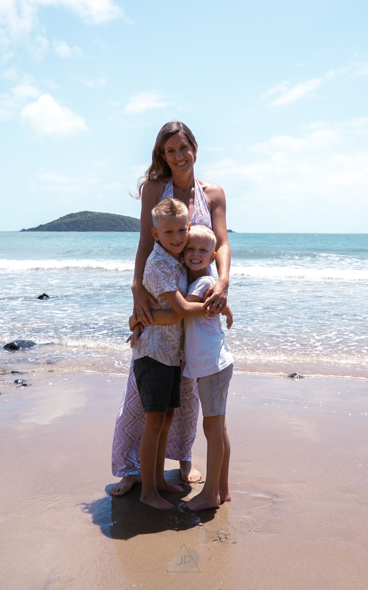 People embracing on a sunny beach with ocean waves and mountains in the background.