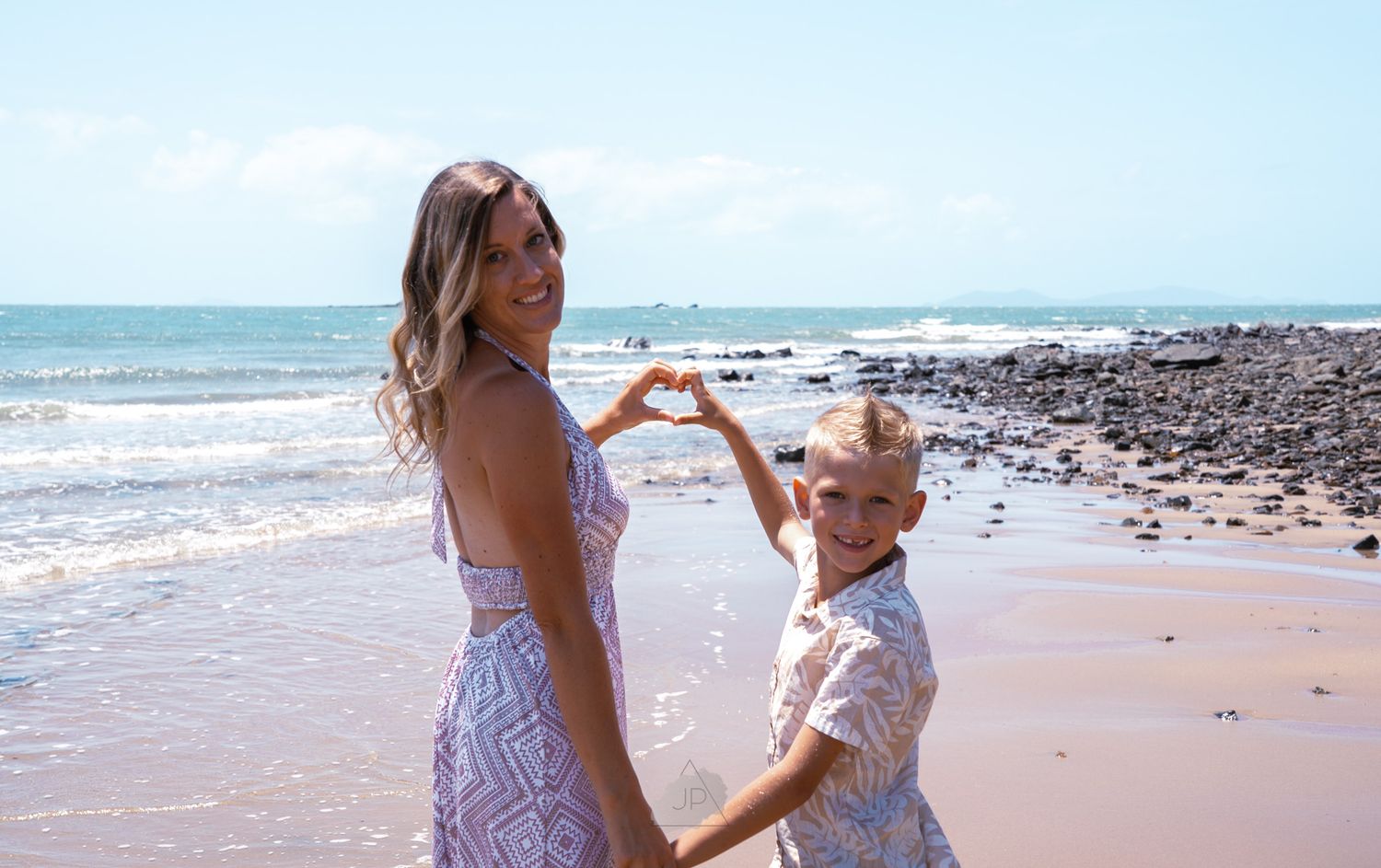 Two people dance and embrace together on a sunny beach with ocean waves in the background.