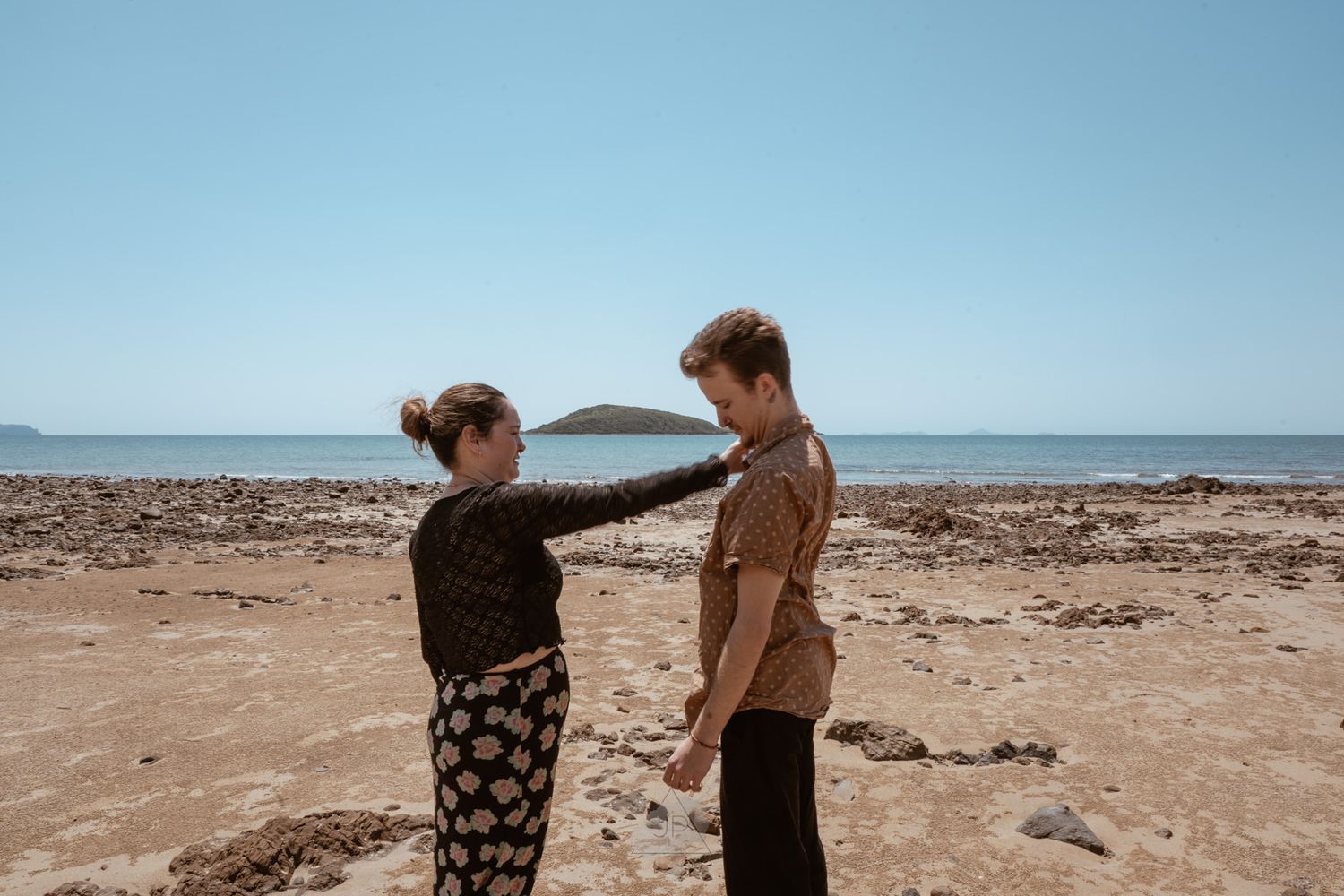 Two people walk along a rocky beach shoreline with waves and blue sky in the background.