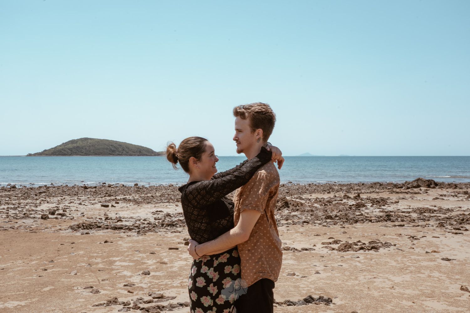 Two people embrace on a rocky beach shoreline with blue ocean waters and clear skies in the background.