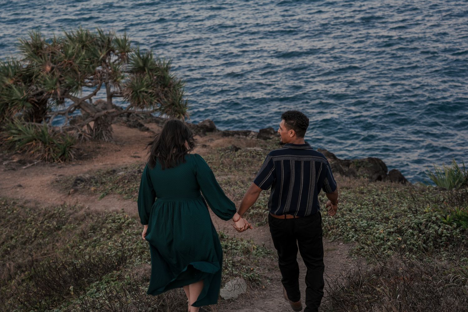 A couple holding hands walks along a scenic coastal cliff path overlooking the blue ocean at dusk.