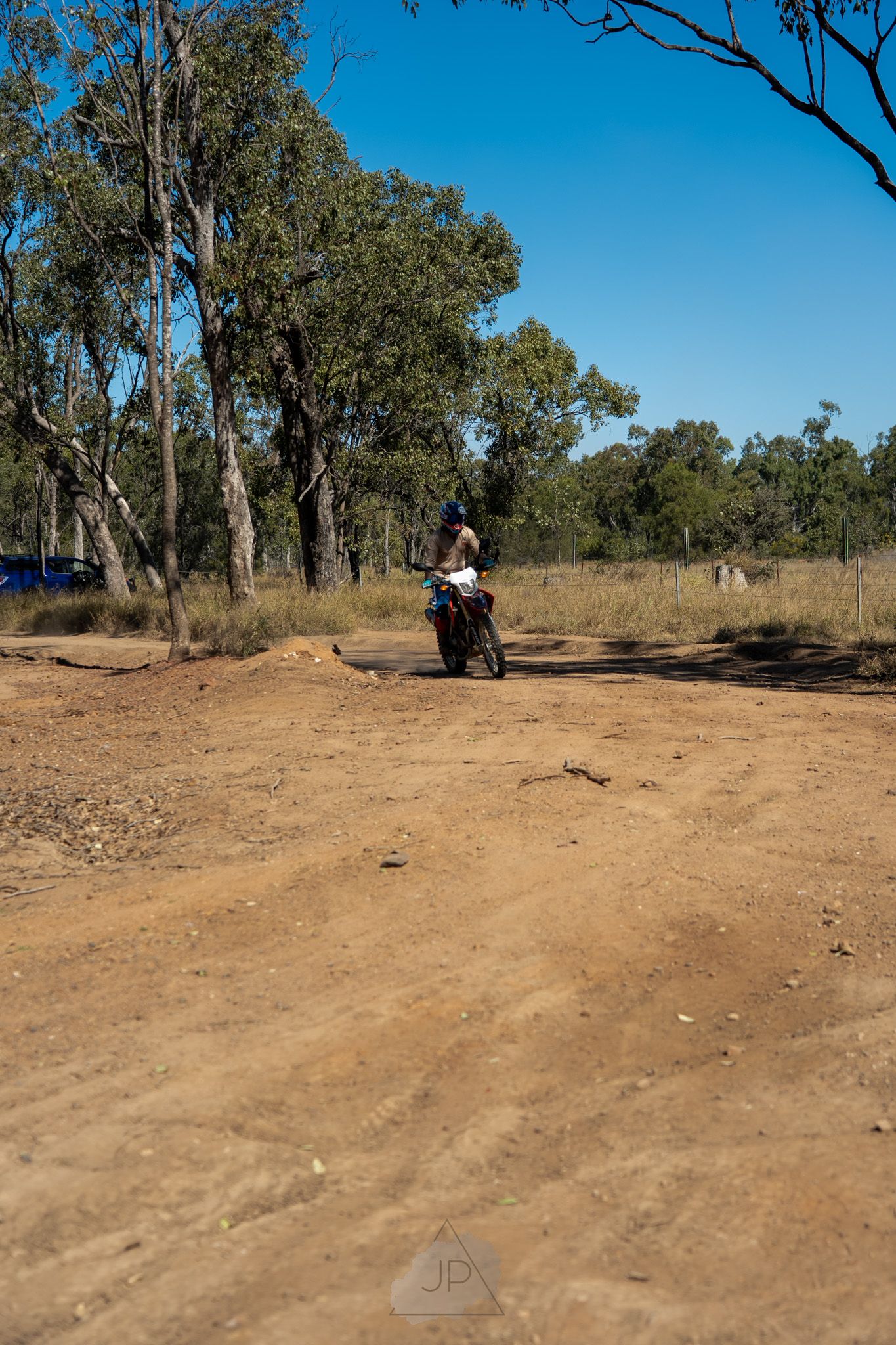 Dirt bike riders navigate through a dusty outback trail lined with eucalyptus trees under bright blue skies.