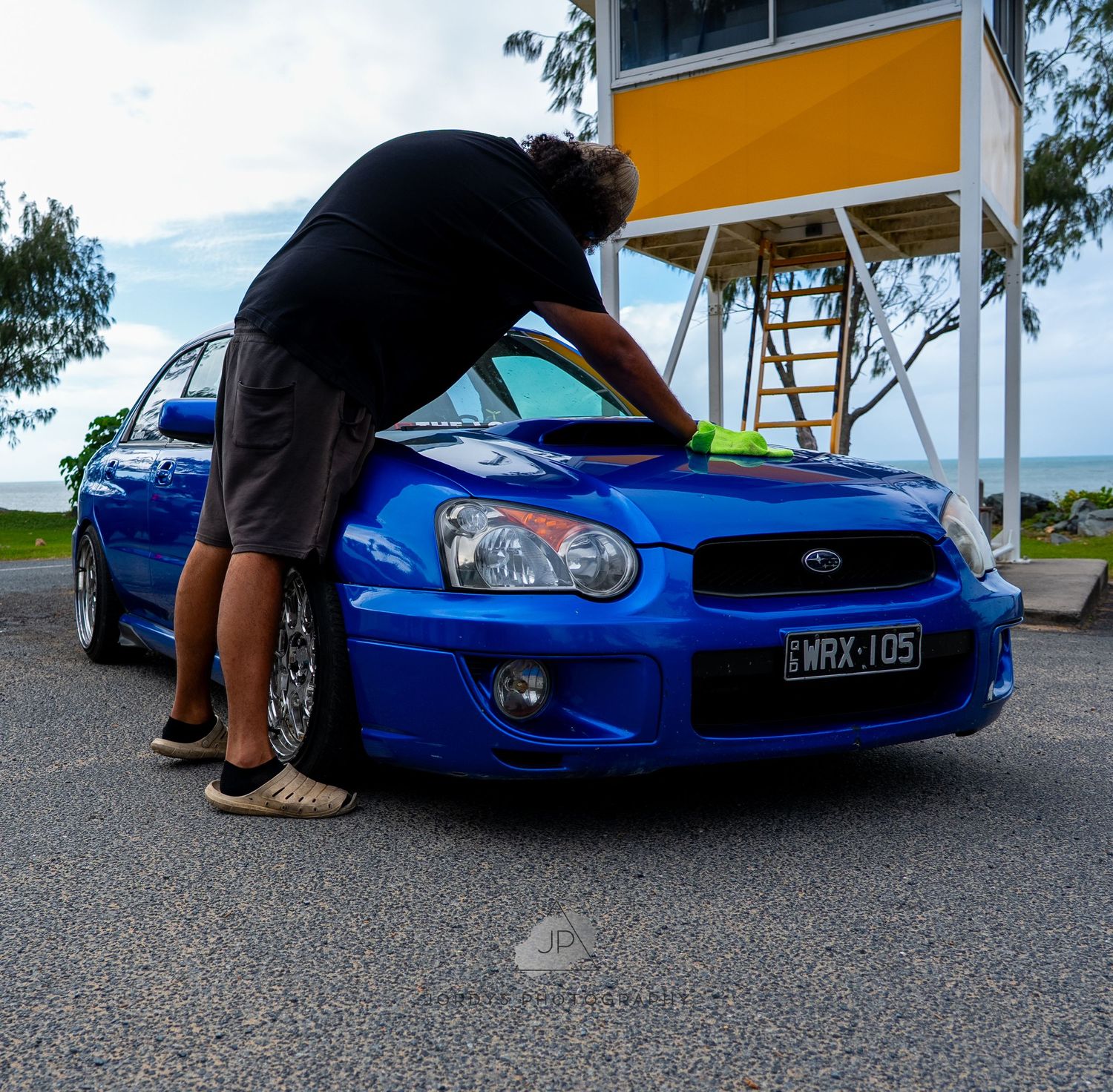 Someone washing a bright blue Subaru WRX on a sunny day at a parking lot with an orange building in the background.