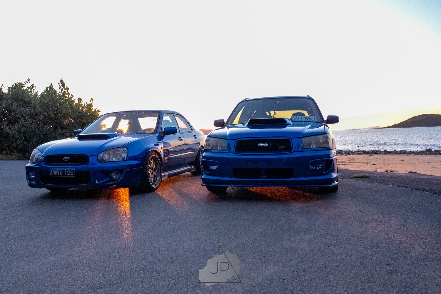 Two blue Subaru WRX STI sports cars parked at sunset with orange underglows illuminating the ground beneath them.