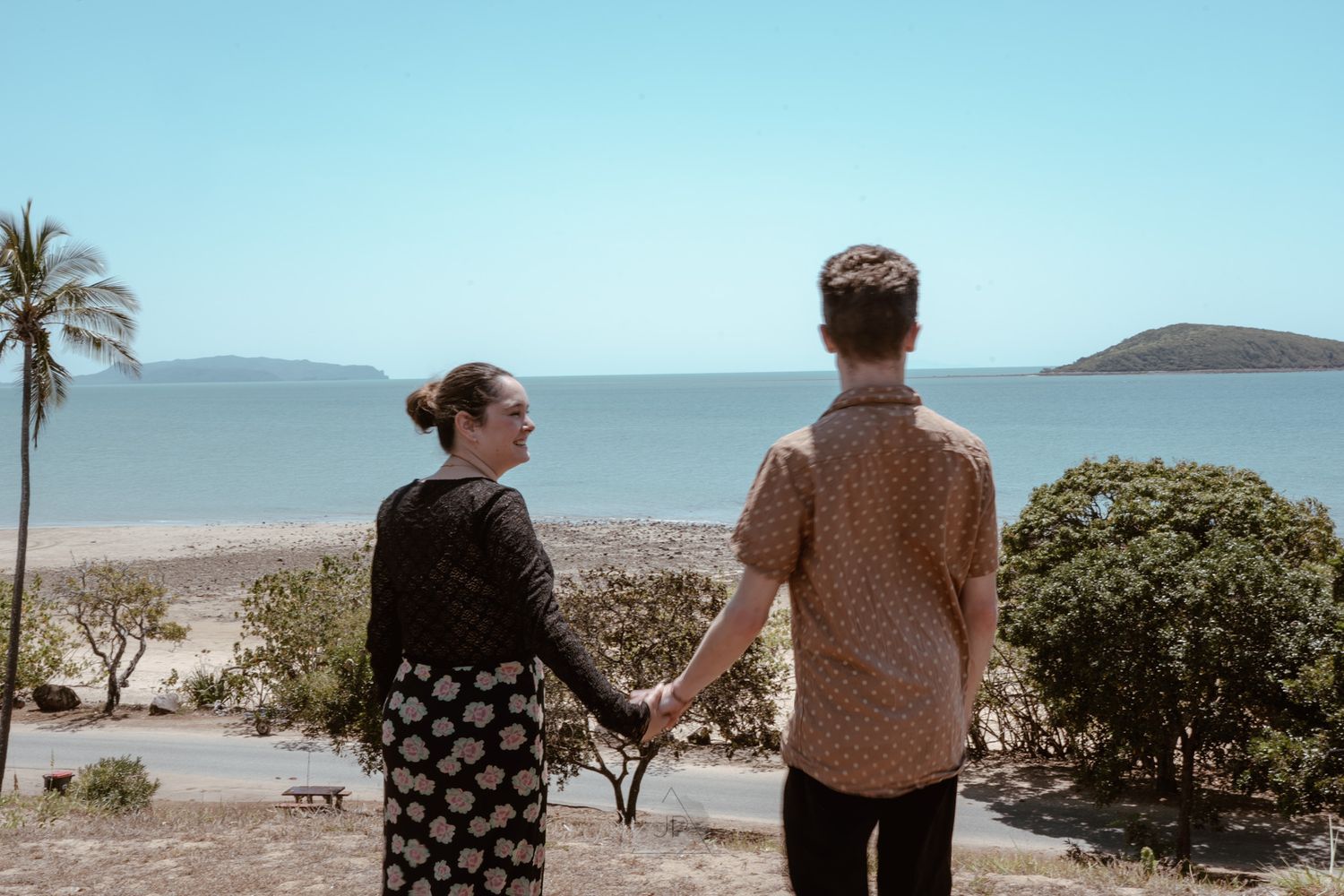 Two people stand together on a coastal overlook with palm trees and ocean views on a sunny day.