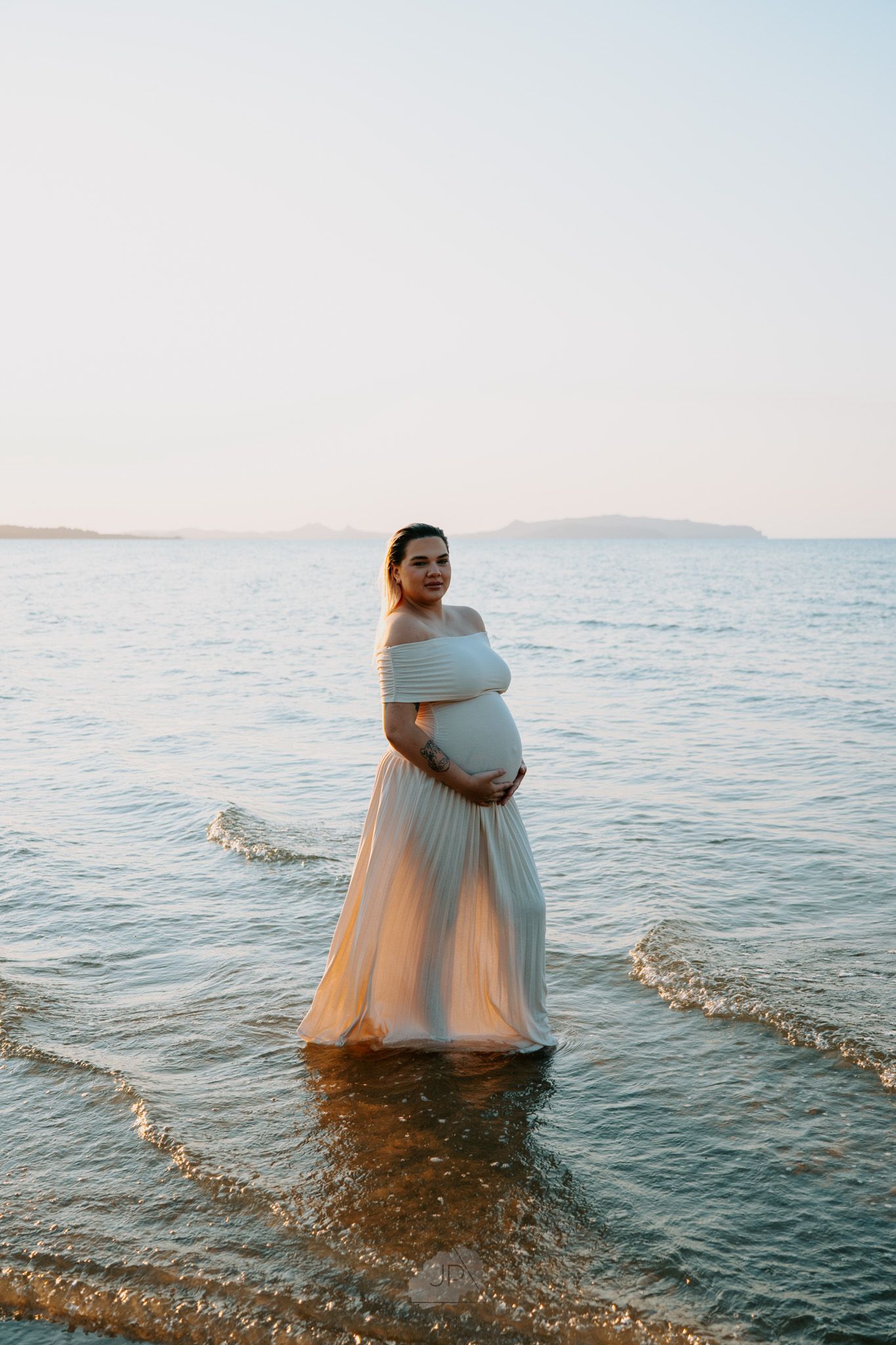 A person in a flowing white dress stands in shallow ocean water during sunset creating a dreamy coastal scene.