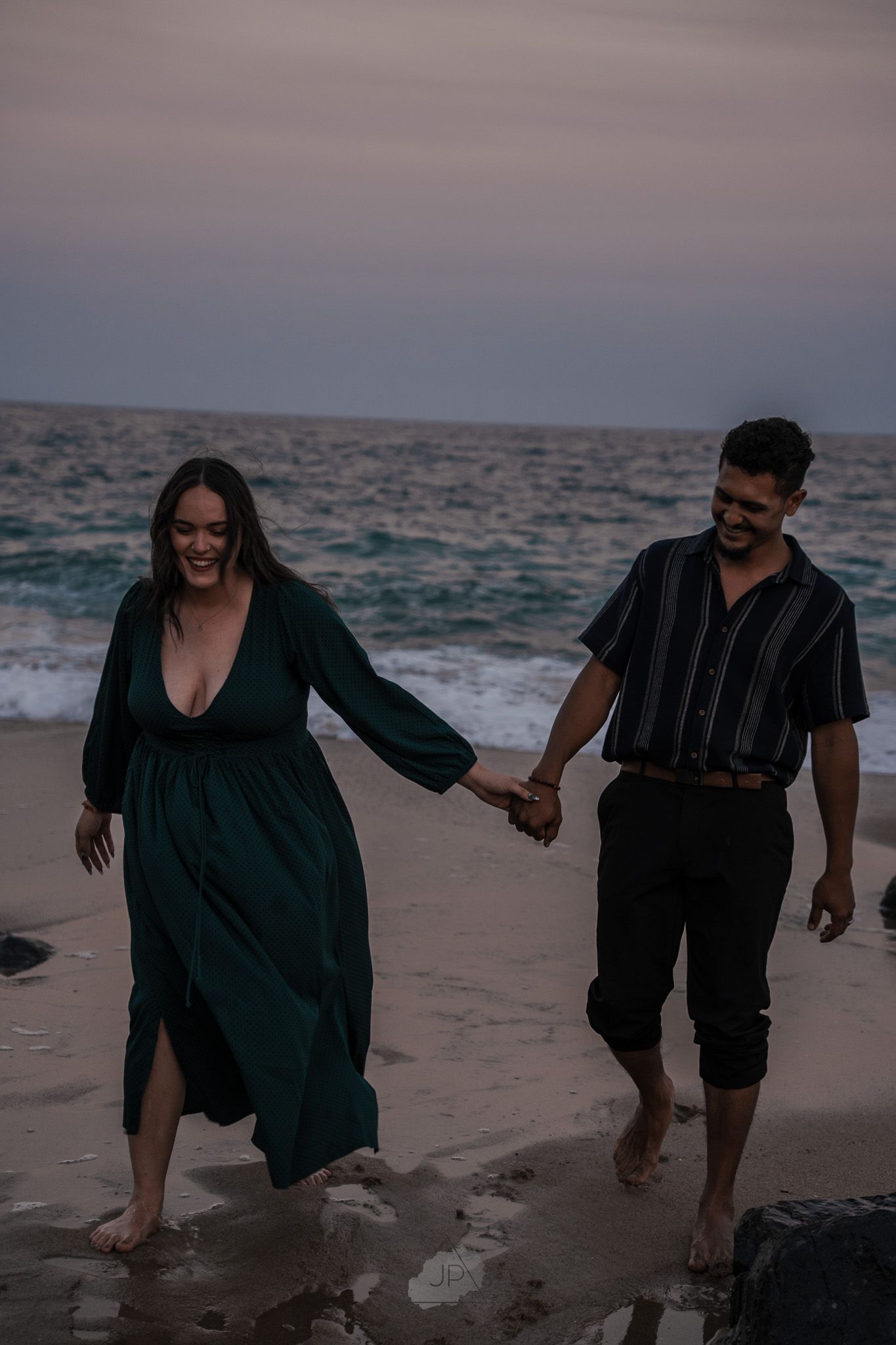 Two people stroll together on a misty beach during sunset, their dark figures contrasting against the grey seascape.