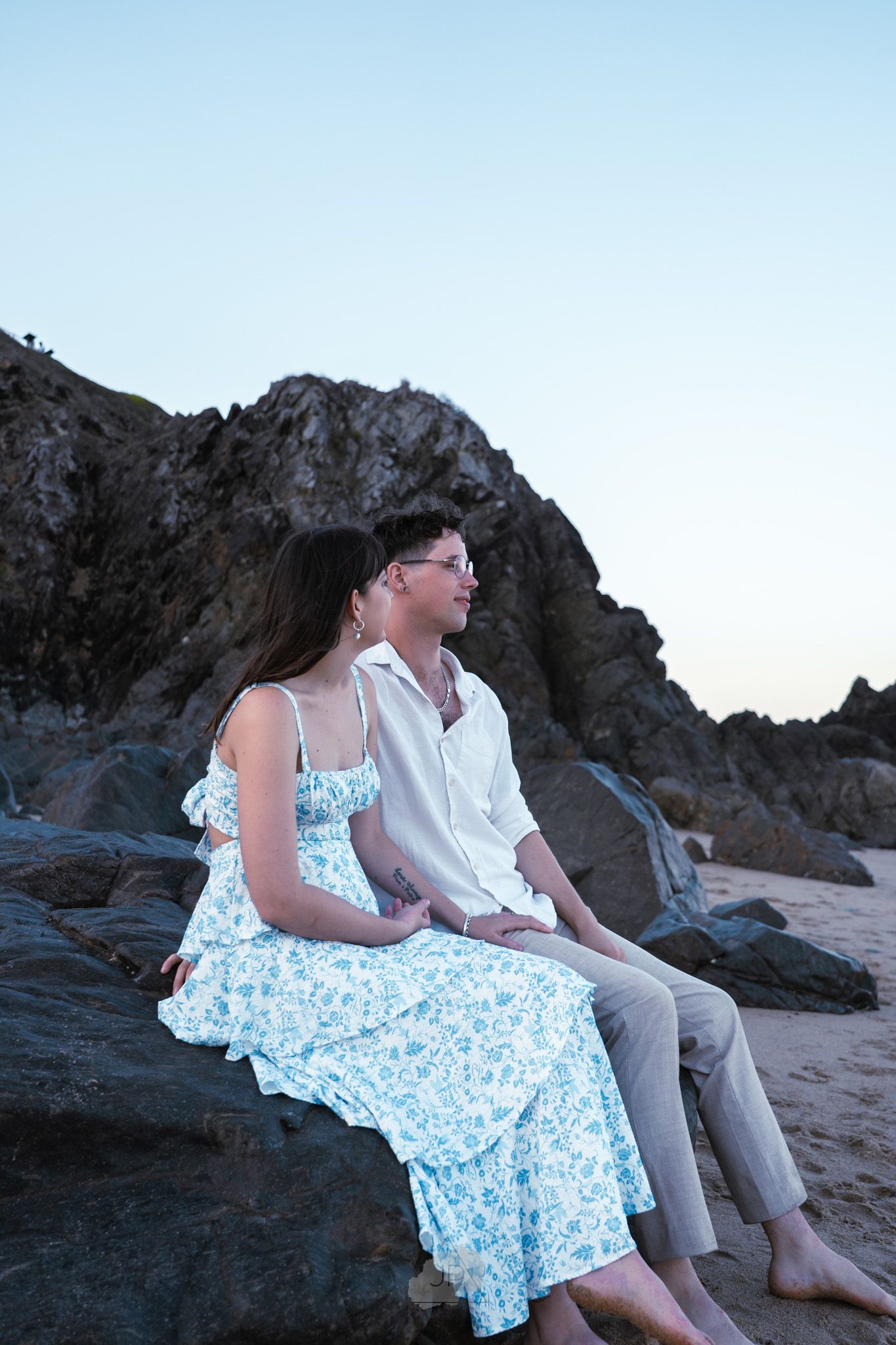 A couple in summer attire sits together on coastal rocks at sunset.