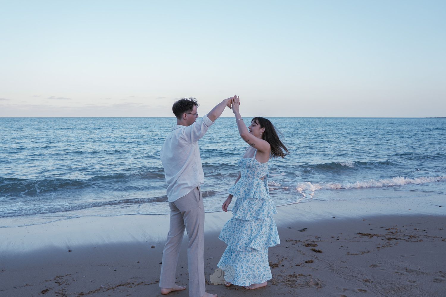 A couple in white and blue outfits dance together on a sandy beach at sunset with ocean waves in the background.