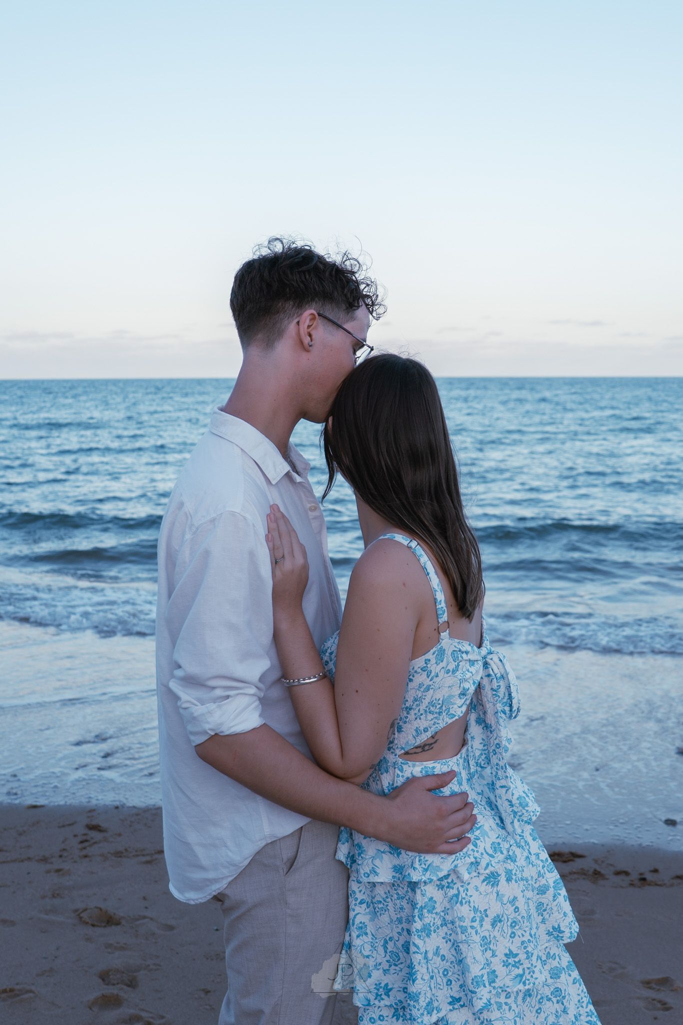 A romantic couple embraces on a sandy beach at sunset with ocean waves in the background.