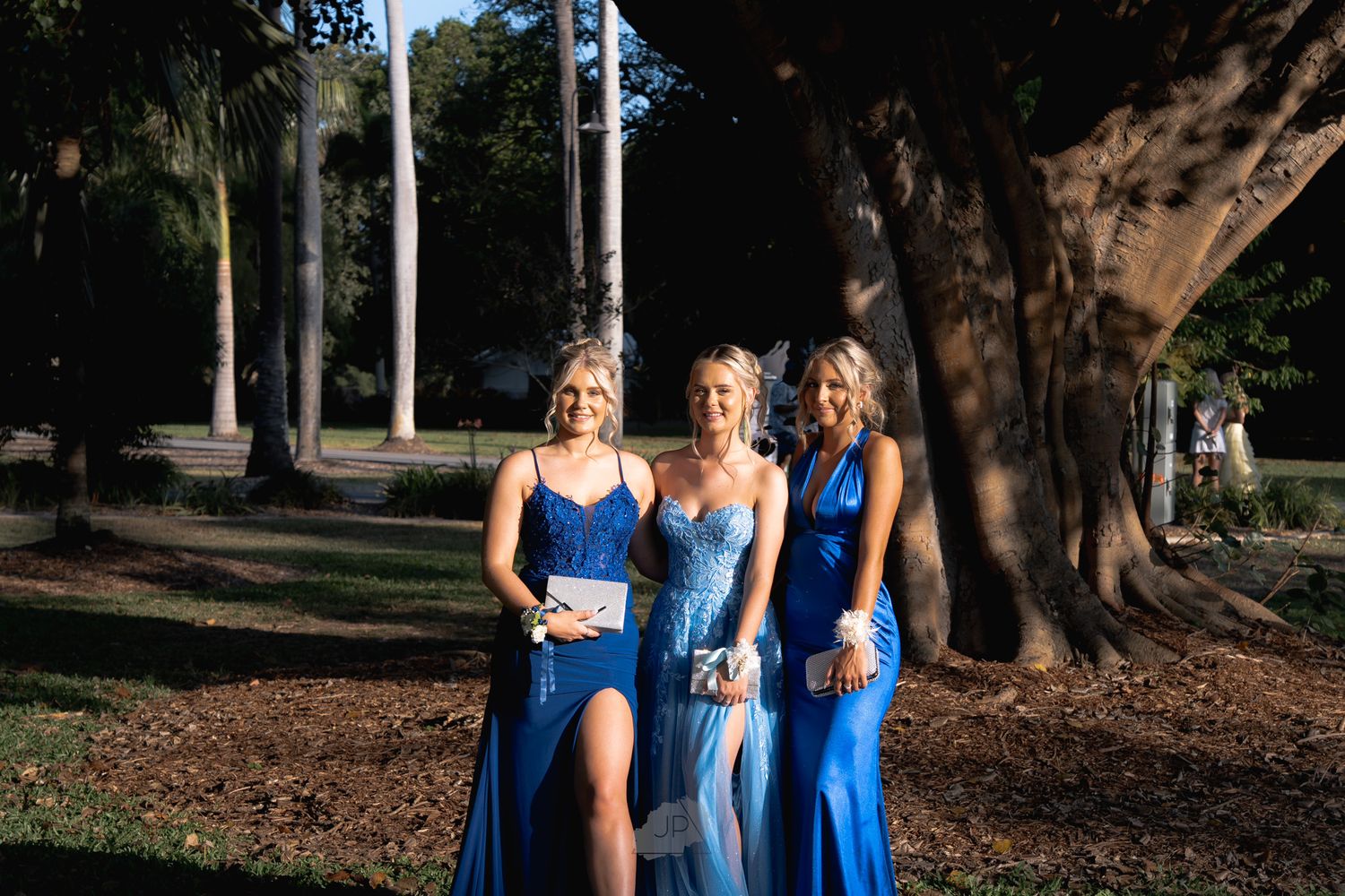 Group wearing matching blue dresses poses together in a sunlit outdoor garden setting.