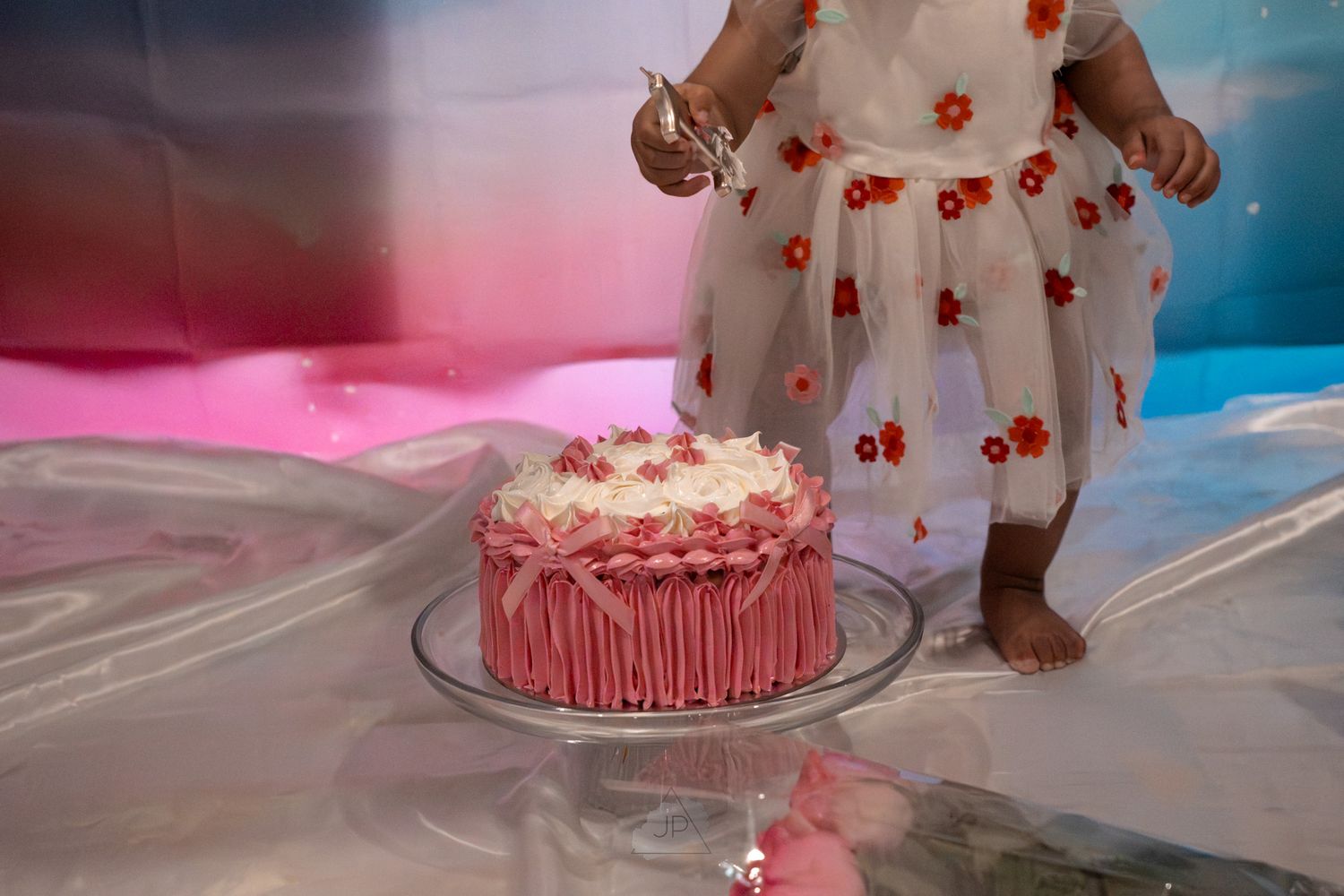 Pink and white birthday cake with delicate floral decorations and ruffled frosting details sits on a display stand.