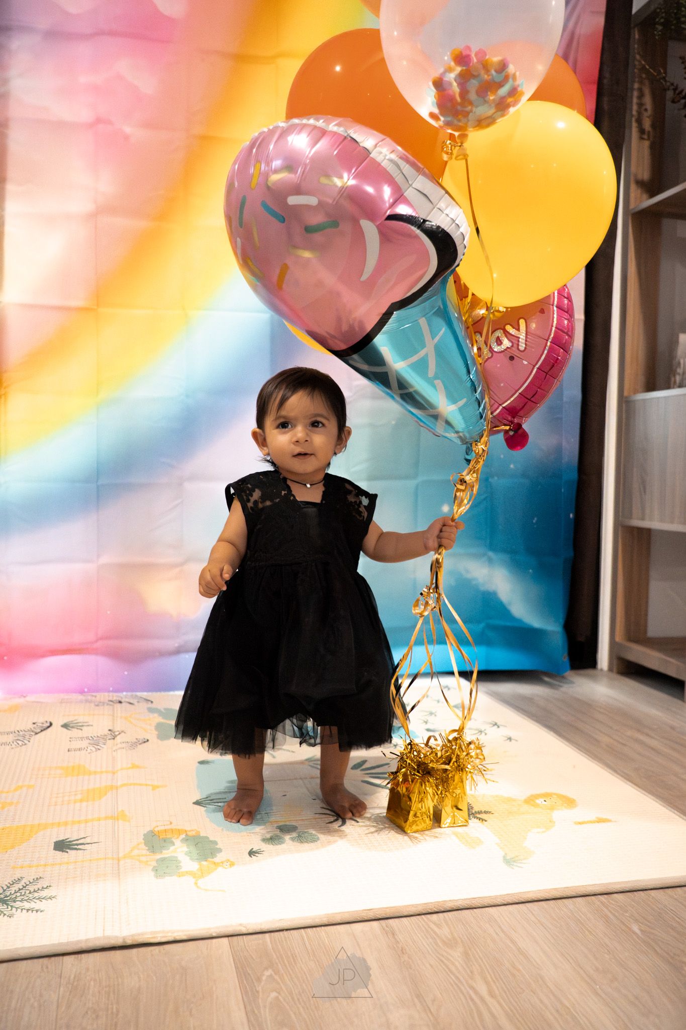 Small child in black dress stands with colorful birthday balloons against rainbow backdrop during celebration.
