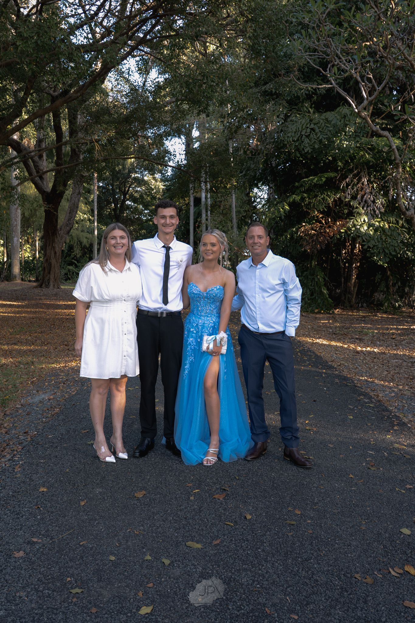 Group of people in formal attire standing outdoors on dark pavement with trees in the background.
