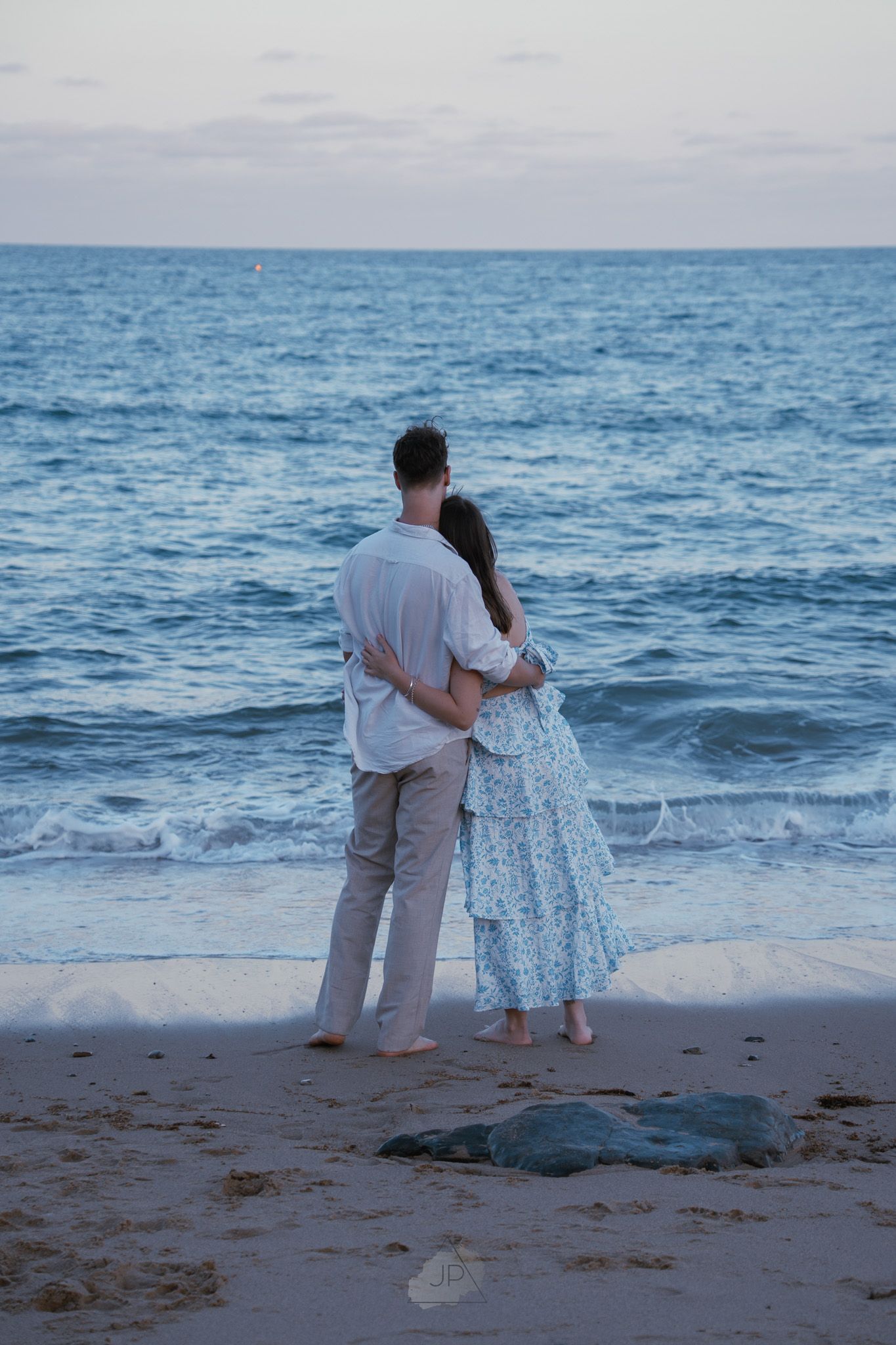 Two people stand together on a beach at dusk watching a sea turtle near the ocean's edge.