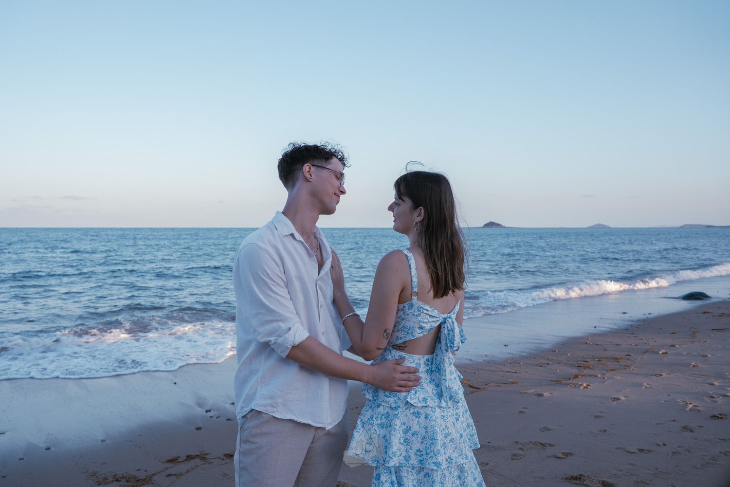Couple in white and blue outfits embracing on a beach at sunset with ocean waves in the background.