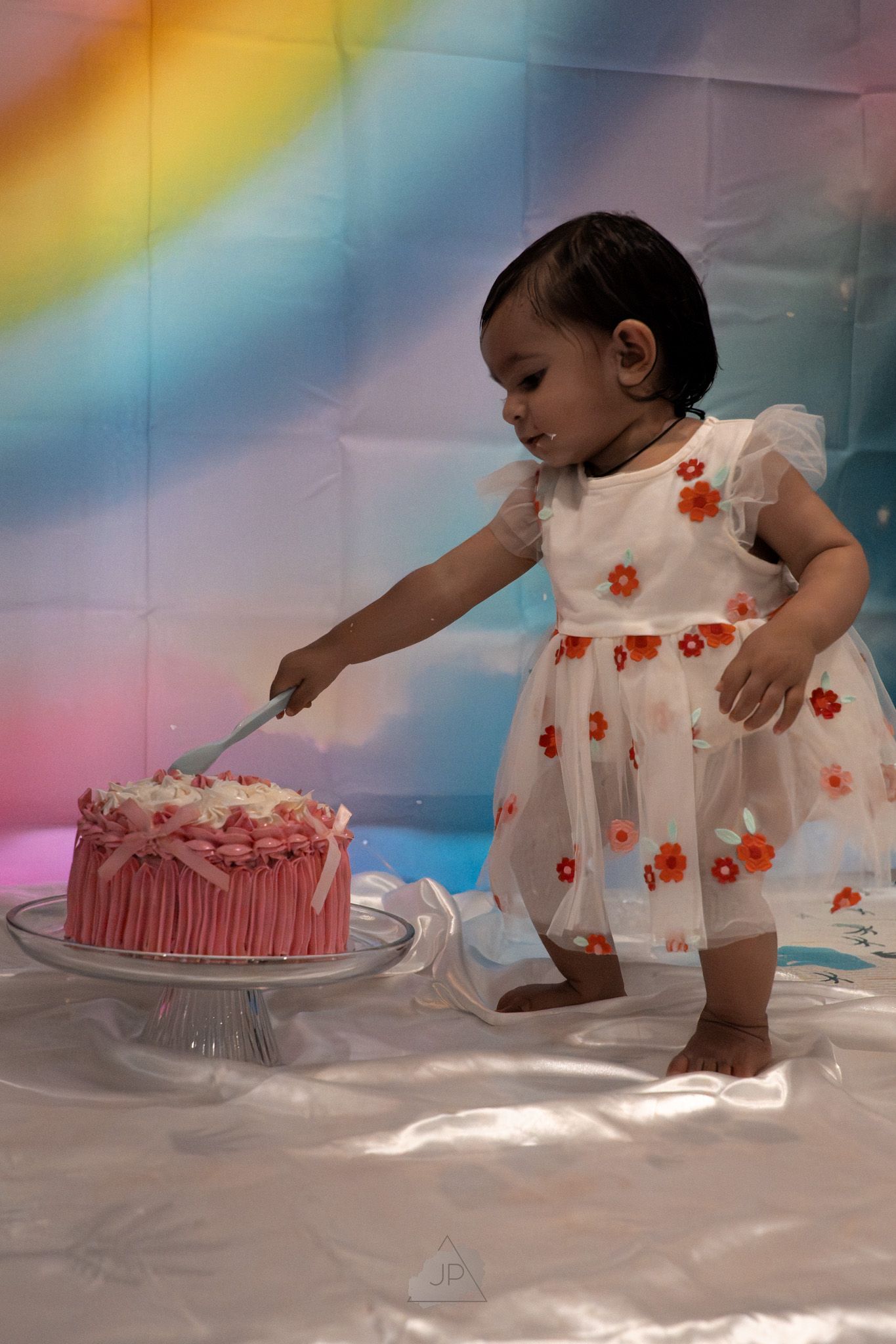 A baby in a white floral dress crawls towards a pink birthday cake in a dreamy, rainbow-lit studio setting.