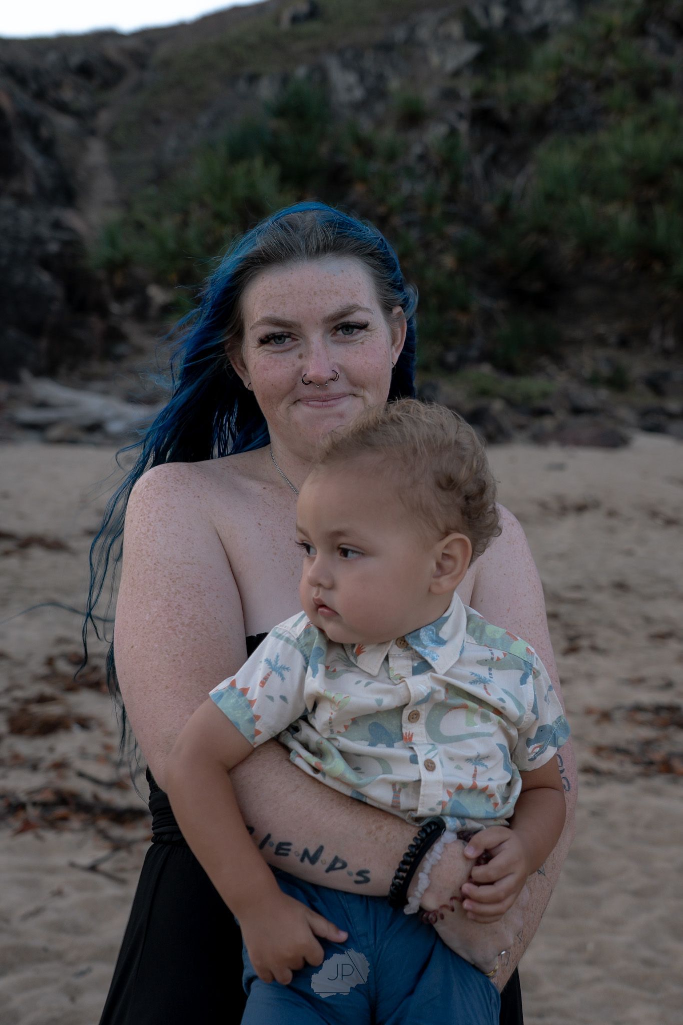 A parent with blue hair holds a toddler wearing a patterned shirt on a beach with cliffs in the background at dusk.