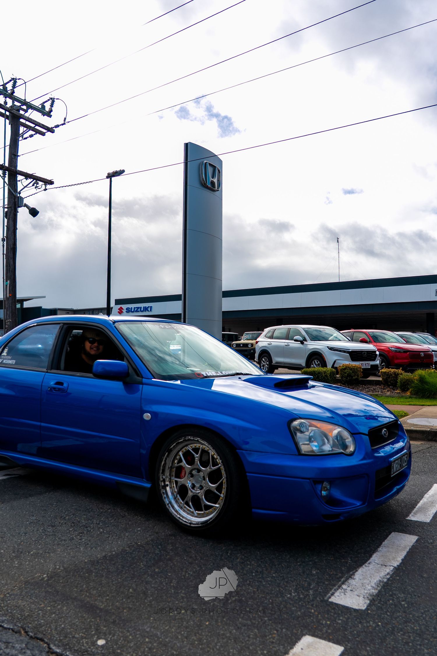 Blue Subaru Impreza WRX STI with aftermarket wheels parked in a lot under cloudy skies.
