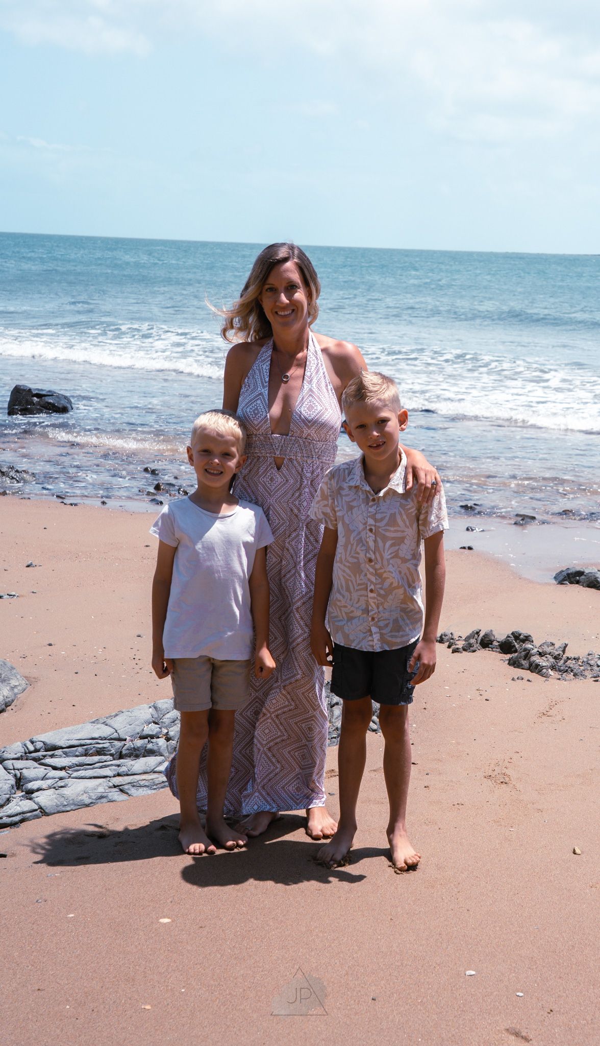 Three people stand together smiling on a sunny beach with blue ocean waters and dark rocks visible behind them.