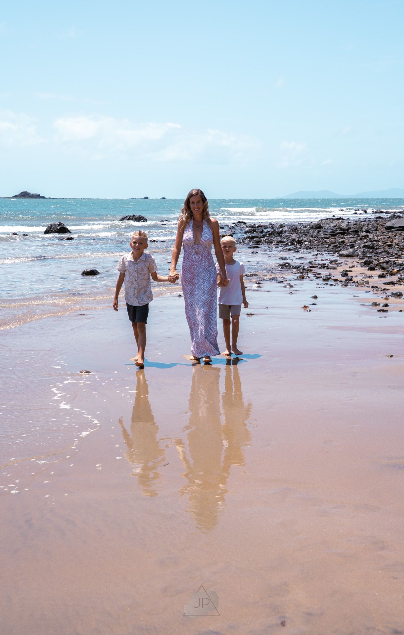 Three people walking together on a scenic beach with rocks and waves, their reflections mirrored in the wet sand.