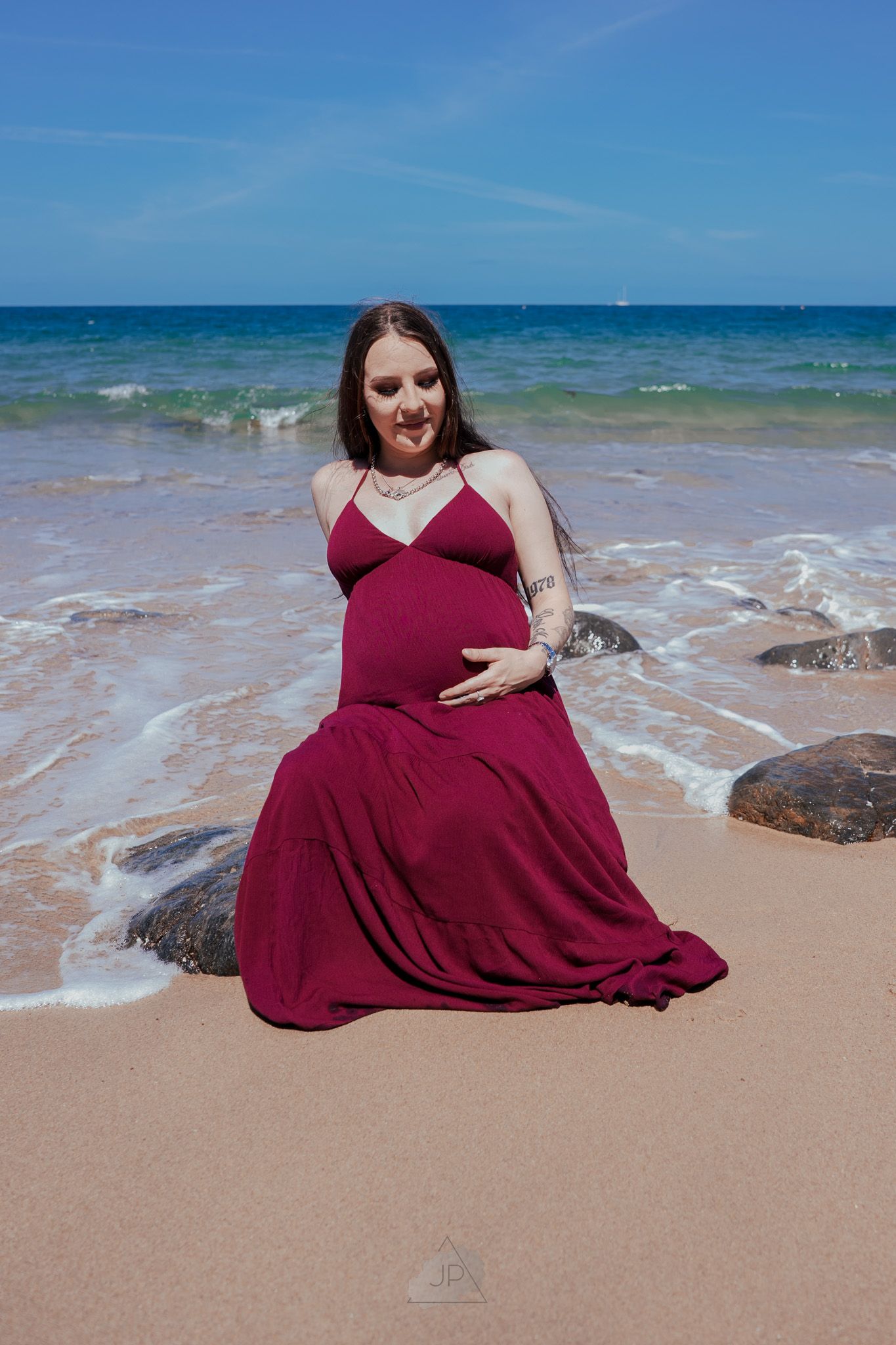 A person in a burgundy dress poses gracefully on a sandy beach with ocean waves and blue sky in the background.