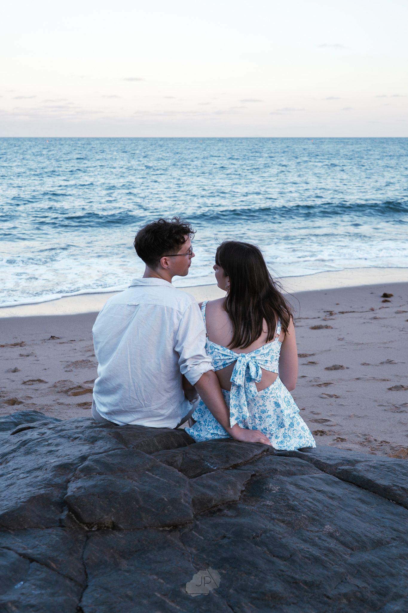 A couple sits together on coastal rocks at the beach during sunset, watching the ocean waves.