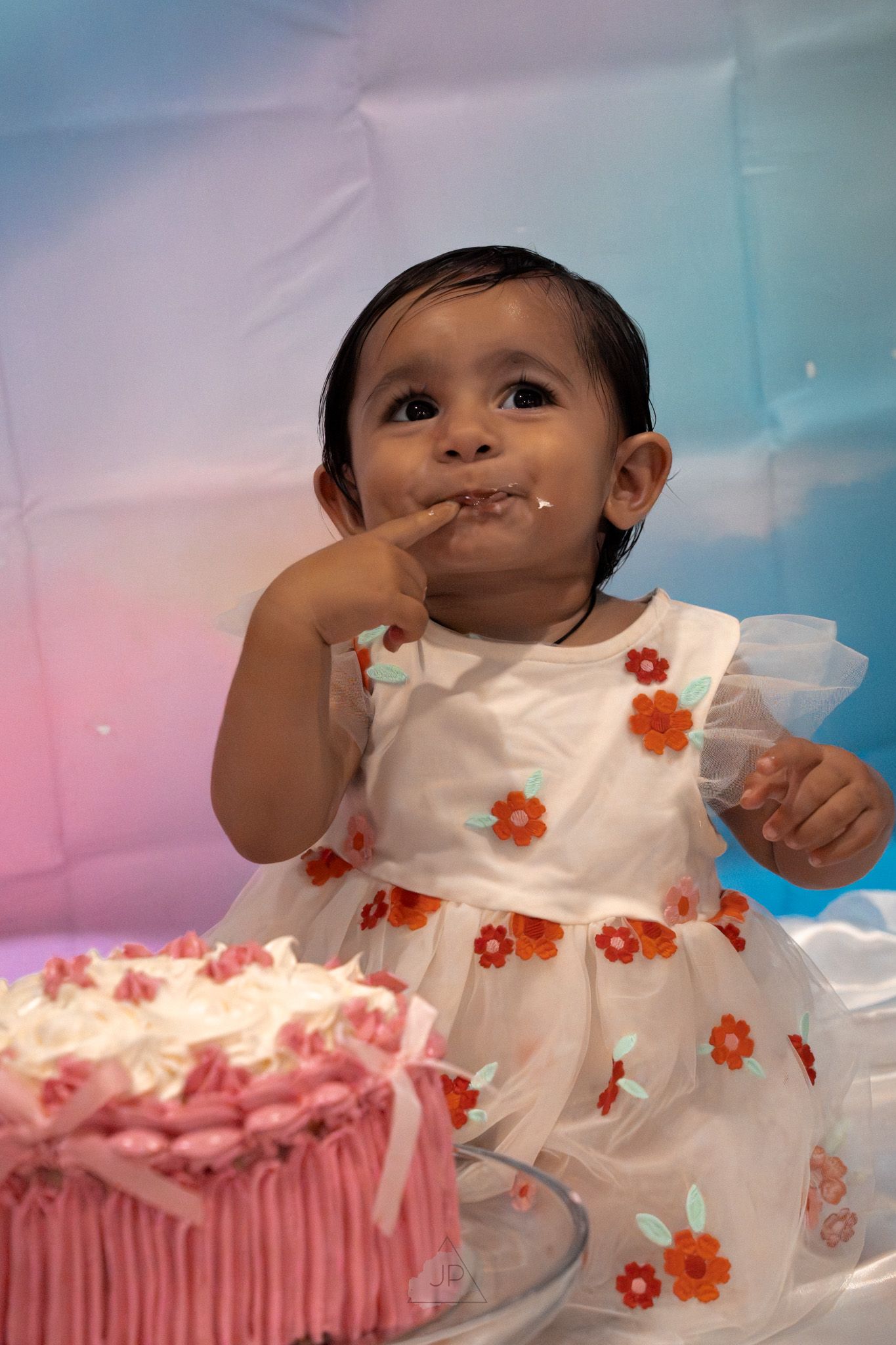 A toddler in a white floral dress tastes frosting from a pink birthday cake during a celebration.