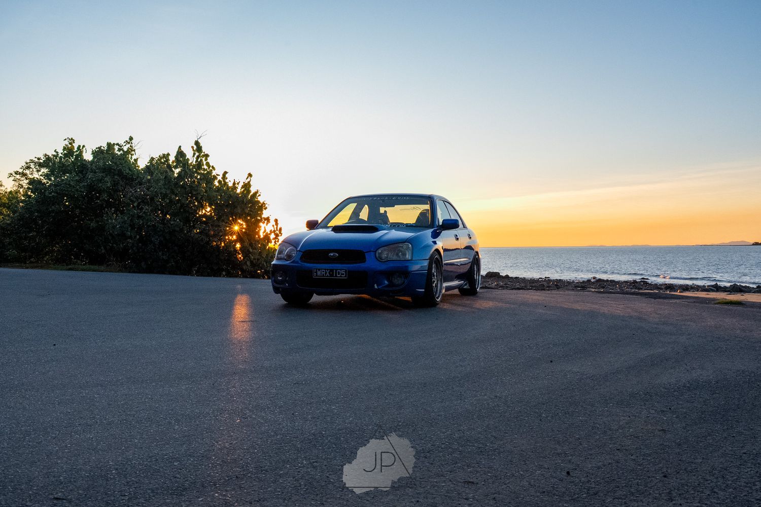 A blue performance car parked on a scenic coastal road at sunset with ocean views in the background.