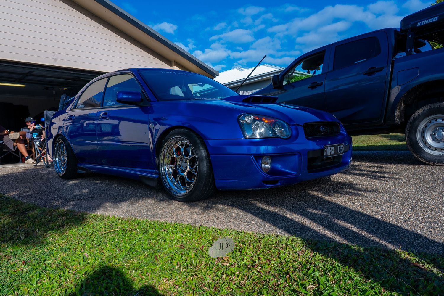 A bright blue modified sports car with custom wheels is parked on a driveway under blue skies.