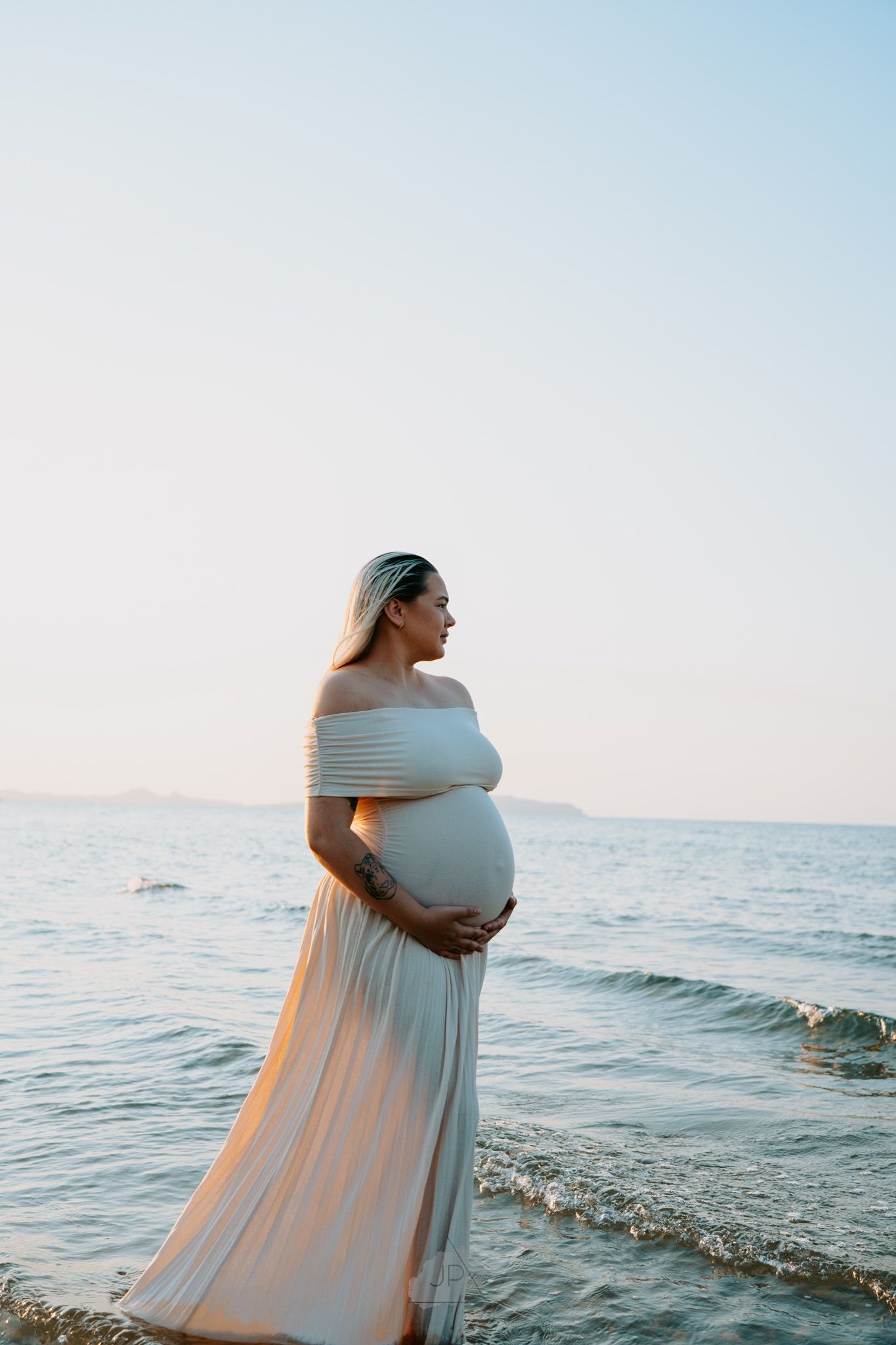 A person in a flowing white dress stands at the ocean's edge during sunset, creating a serene coastal scene.