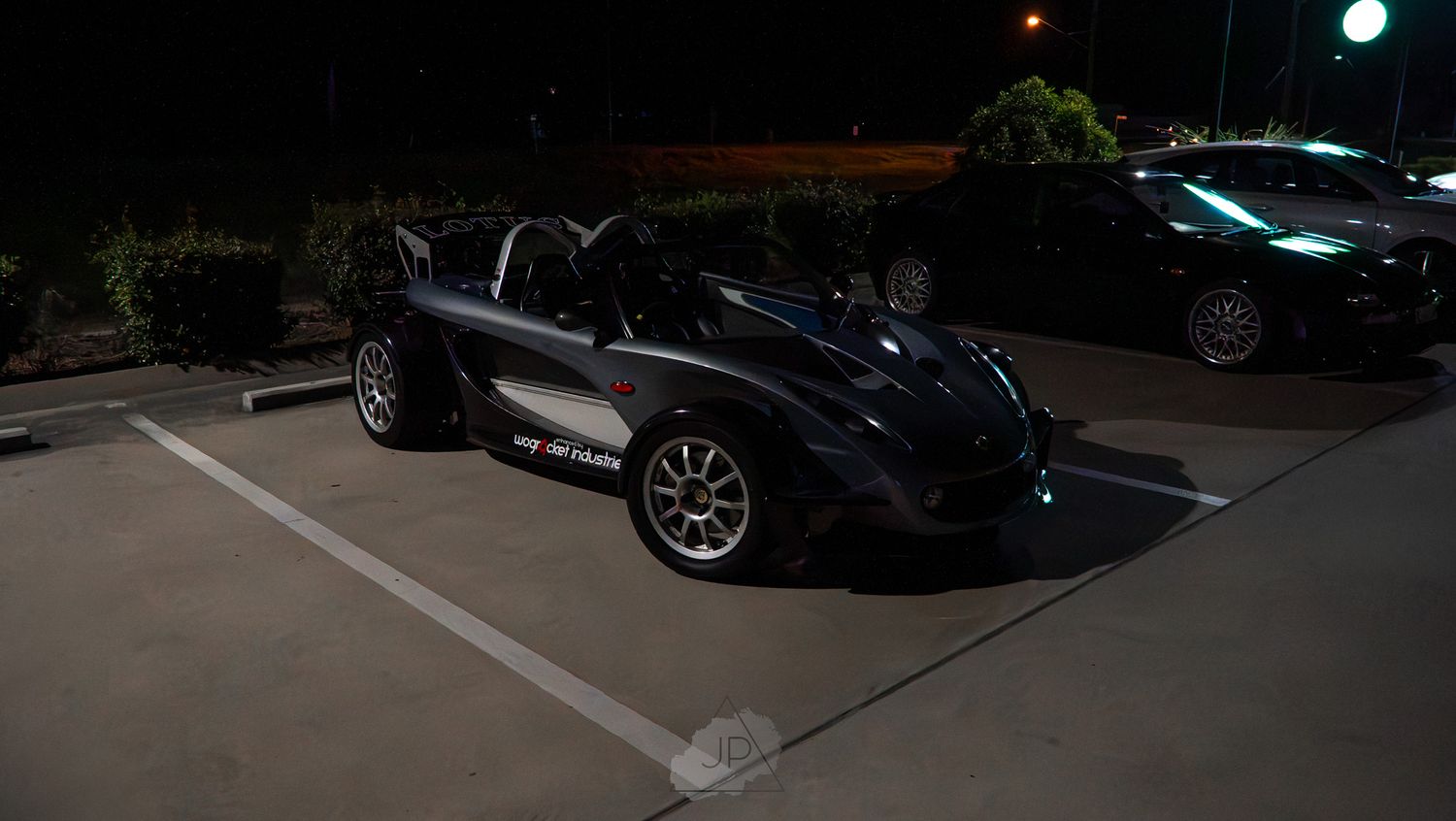 Dark vehicles parked in a dimly lit parking lot under evening city lights next to a brick building.
