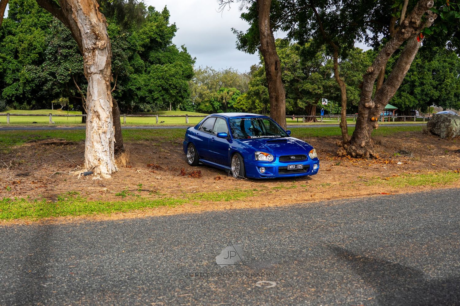 A bright blue Subaru WRX STI parked under tall trees on a curved road.