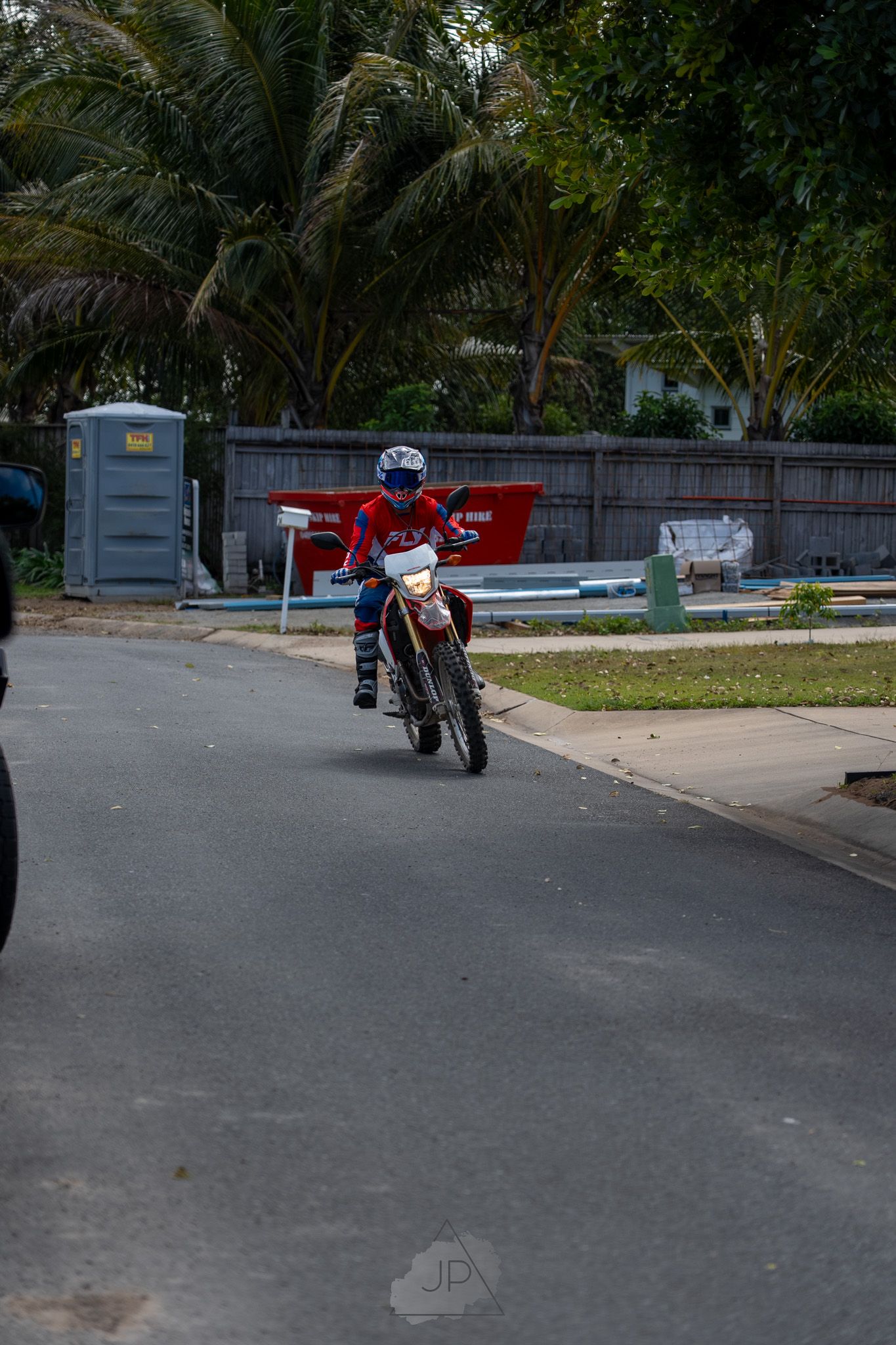 A motorcyclist rides a dirt bike on a residential street lined with palm trees and a portable toilet in the background.