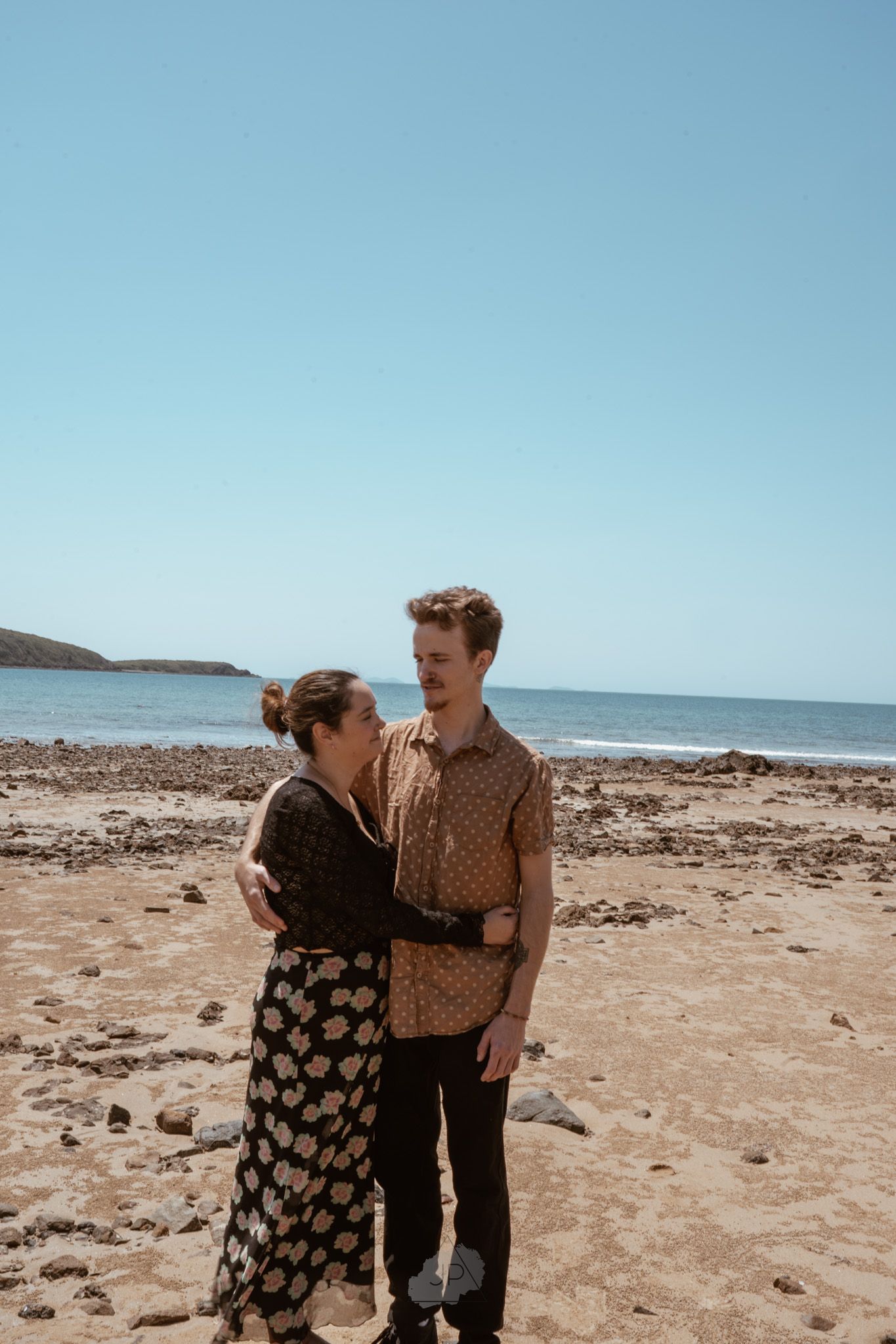 A couple embraces while standing on a rocky beach shoreline with ocean views on a sunny day.