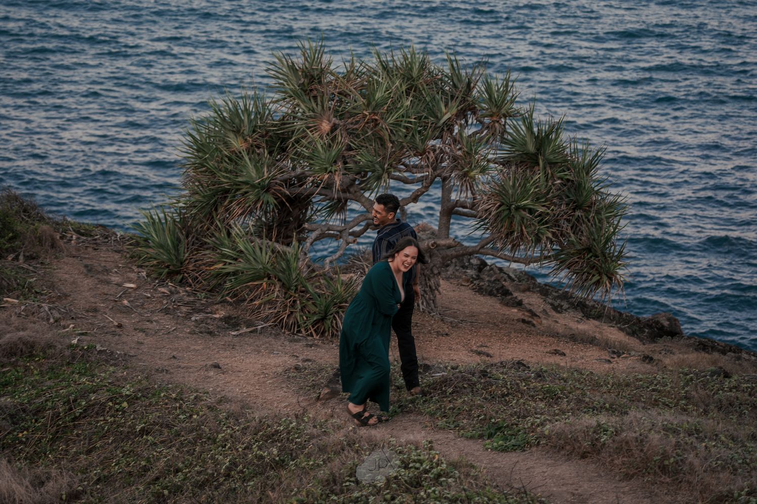 A couple embraces while standing on a coastal cliff with ocean views and native plants surrounding them.