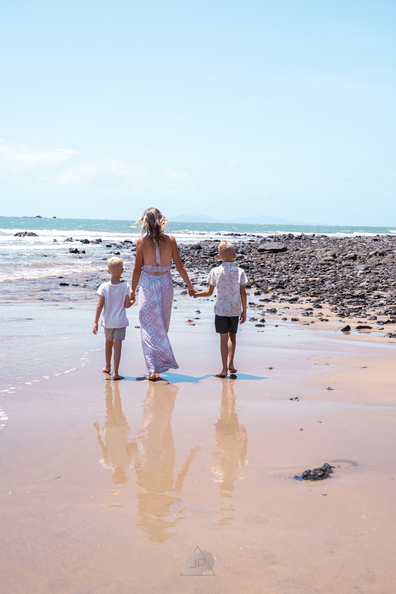 Three people walking along a sandy beach with waves and rocky shoreline in the background.