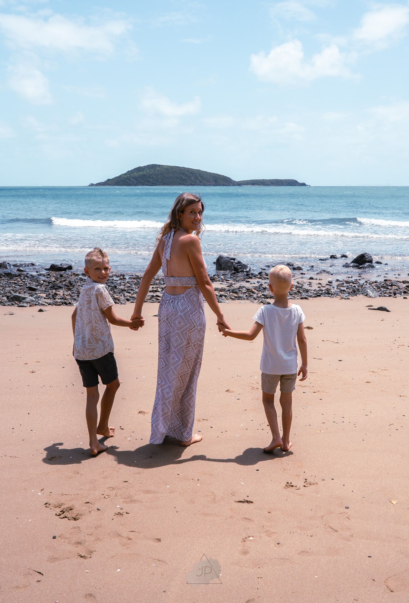 Three figures in white clothing walk together along a scenic beach with an island visible in the background.