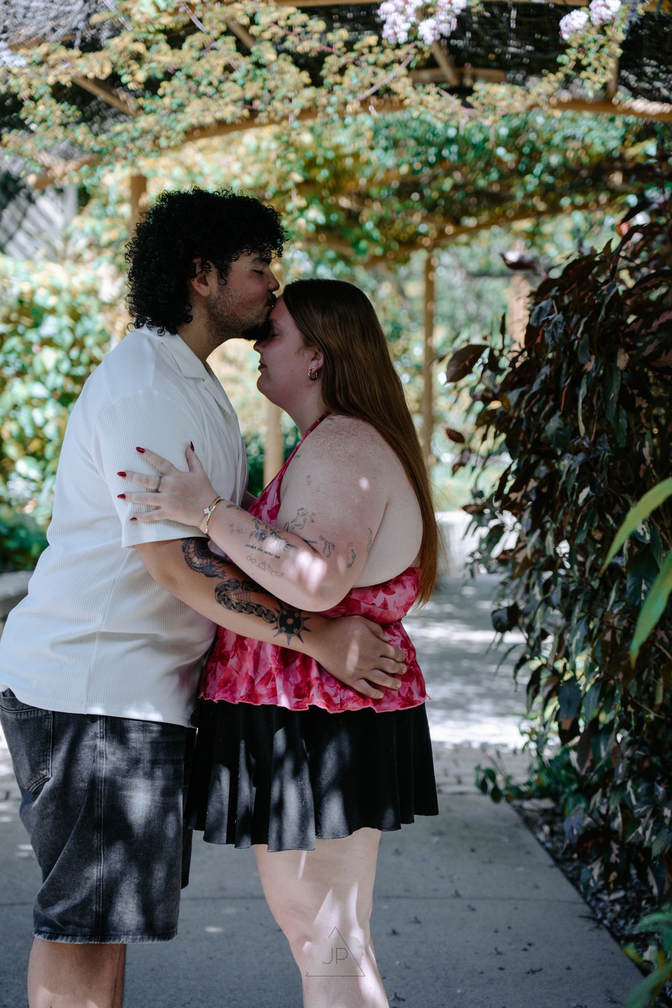 A romantic couple embraces and shares a kiss in a garden pathway with dappled sunlight filtering through trees.