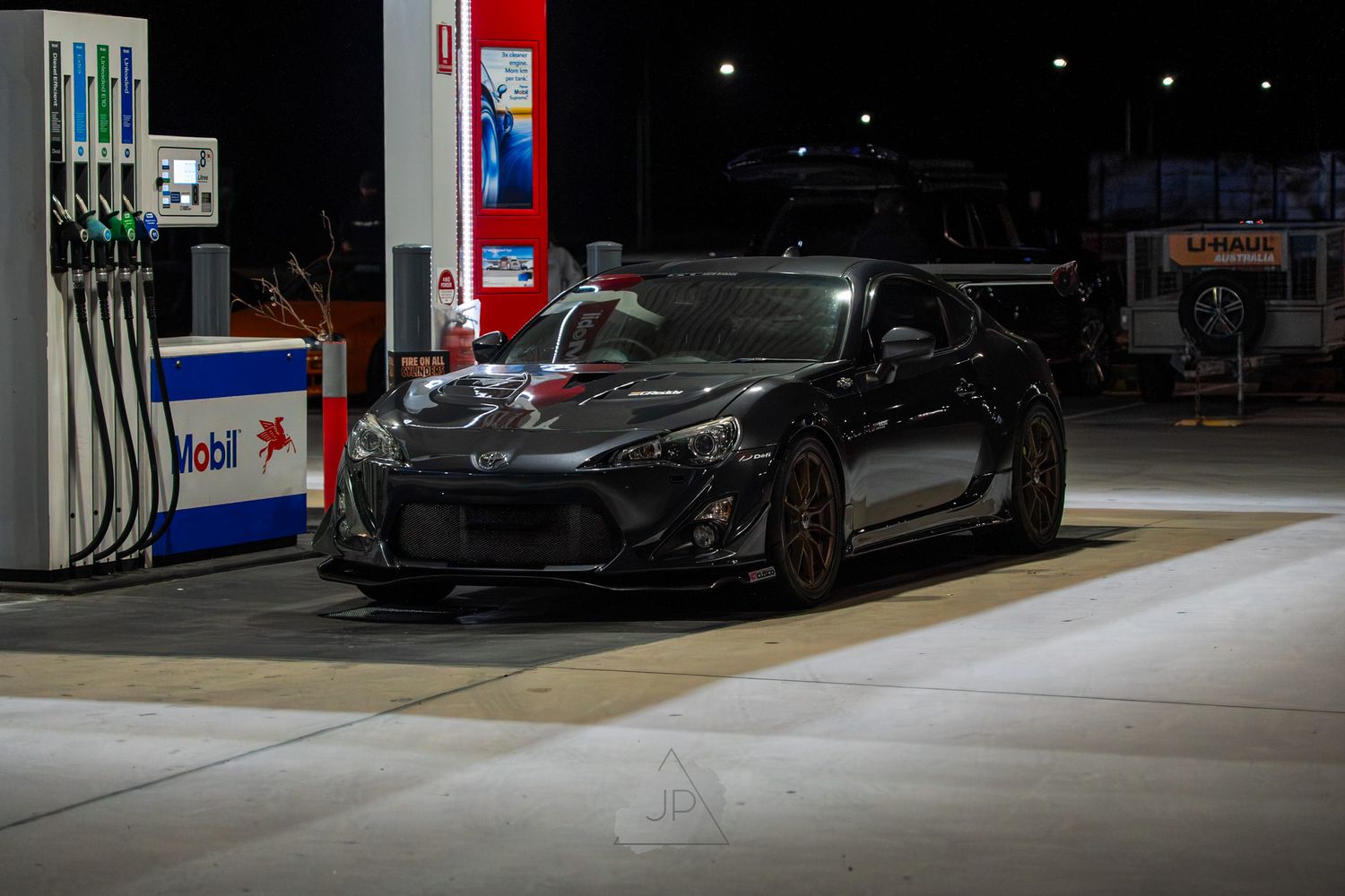 Dark car parked at night under bright lights at a gas station fuel pump.
