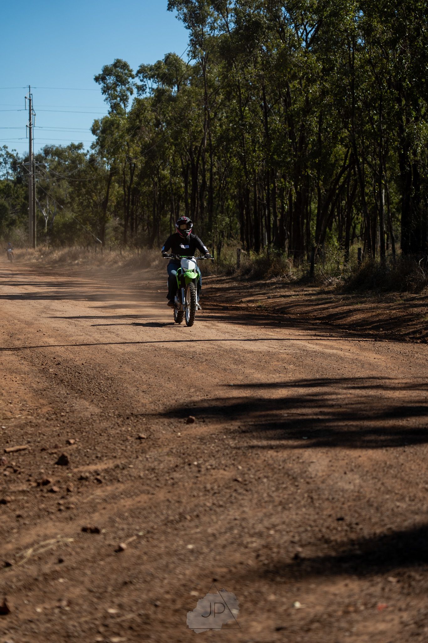 A rugged dirt road winds through an Australian bushland with scattered trees and power lines visible in the distance.