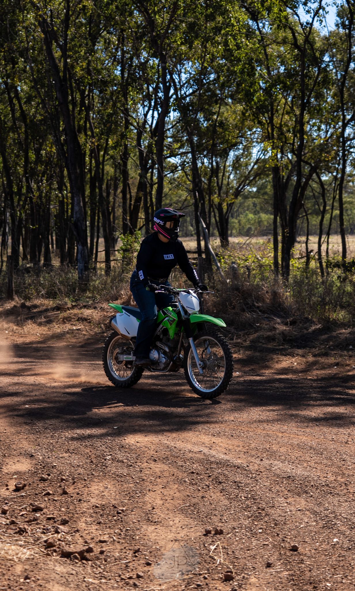 Dirt bike rider navigating through wooded trail on green motorcycle in sunny forest setting.