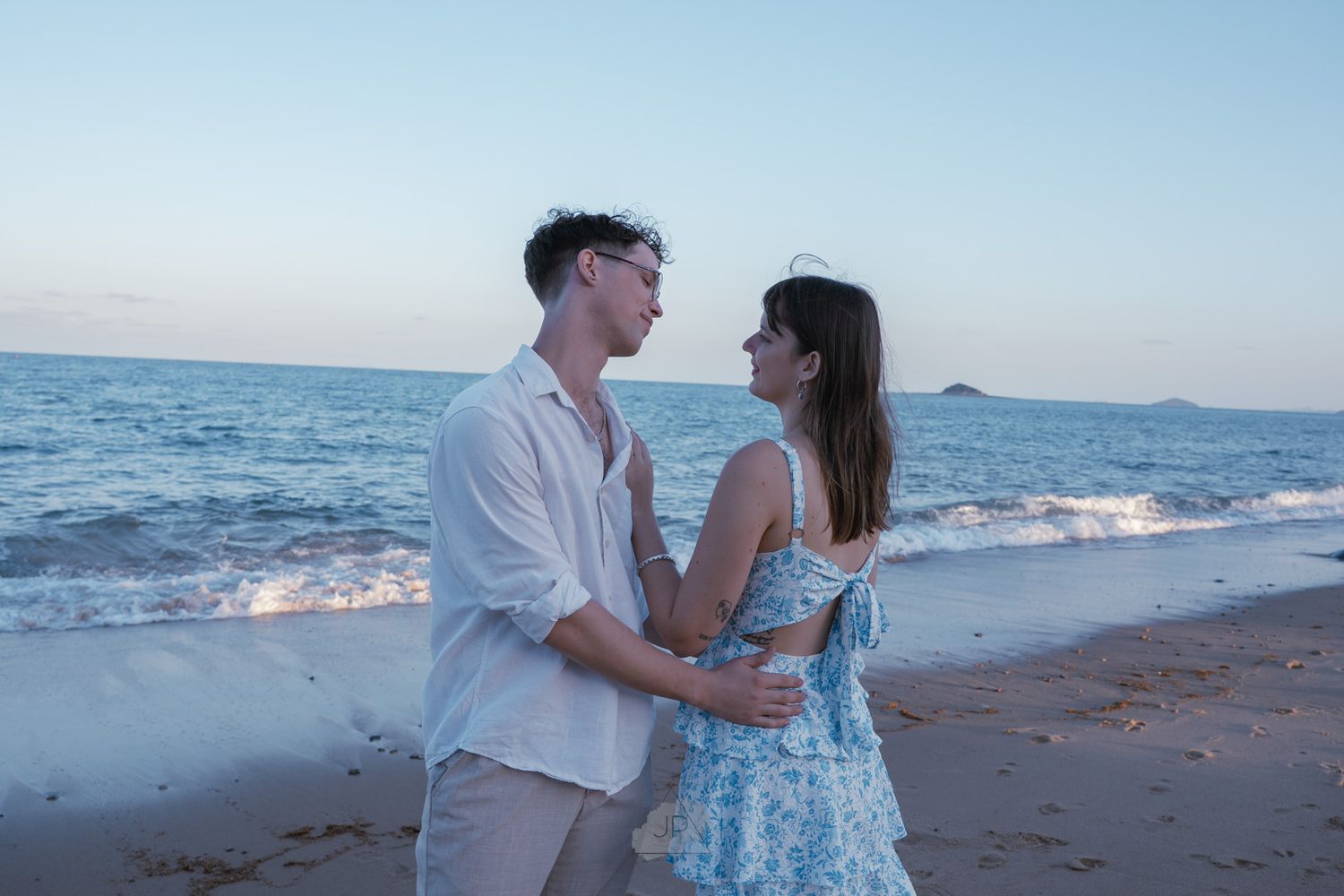 Couple in light blue and white outfits share romantic moments along a scenic beach at sunset.