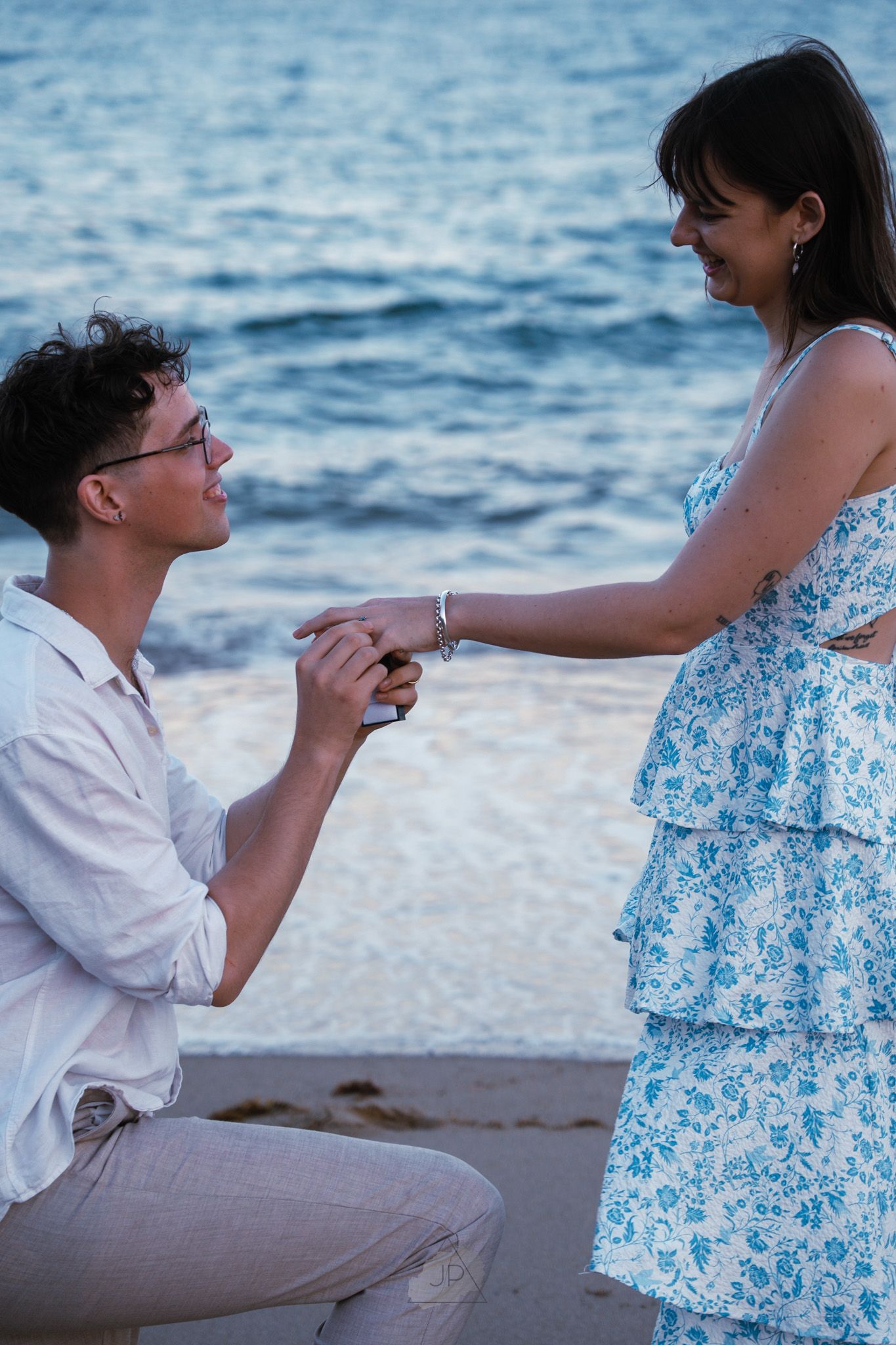 A romantic seaside proposal scene at sunset with a couple in white and blue outfits by the ocean.
