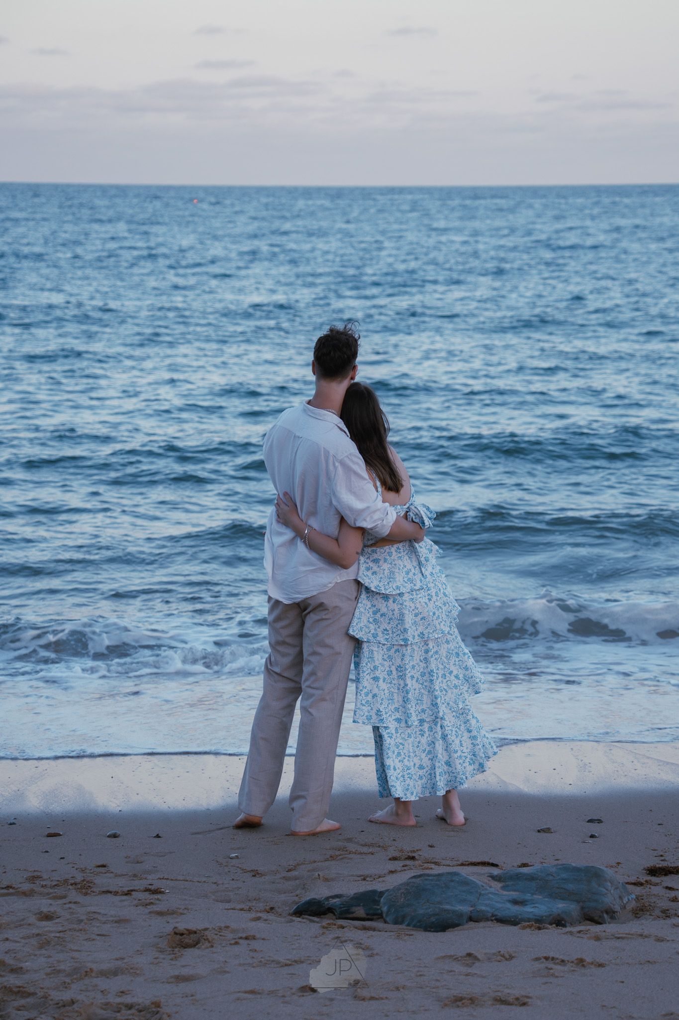 A couple embraces while standing on a beach at dusk overlooking the blue ocean waves.