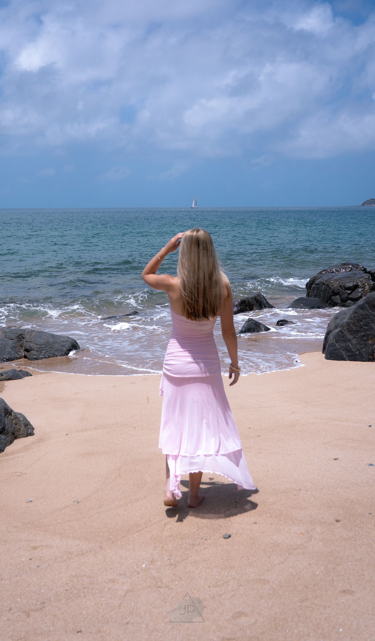 A person in a pink dress stands on a sandy beach near rocky outcrops and ocean waves.