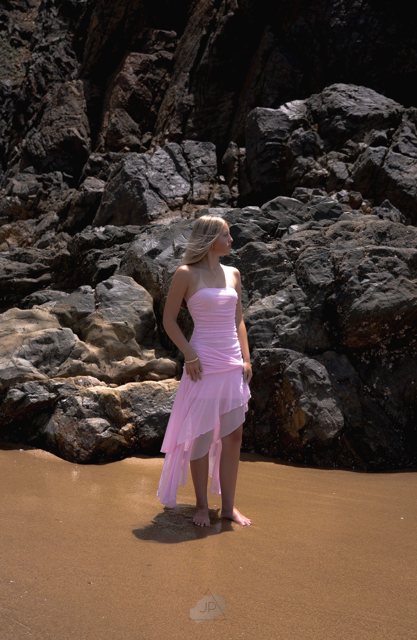 A person in a pink dress stands on a sandy beach against dark rocky cliffs on a sunny day.