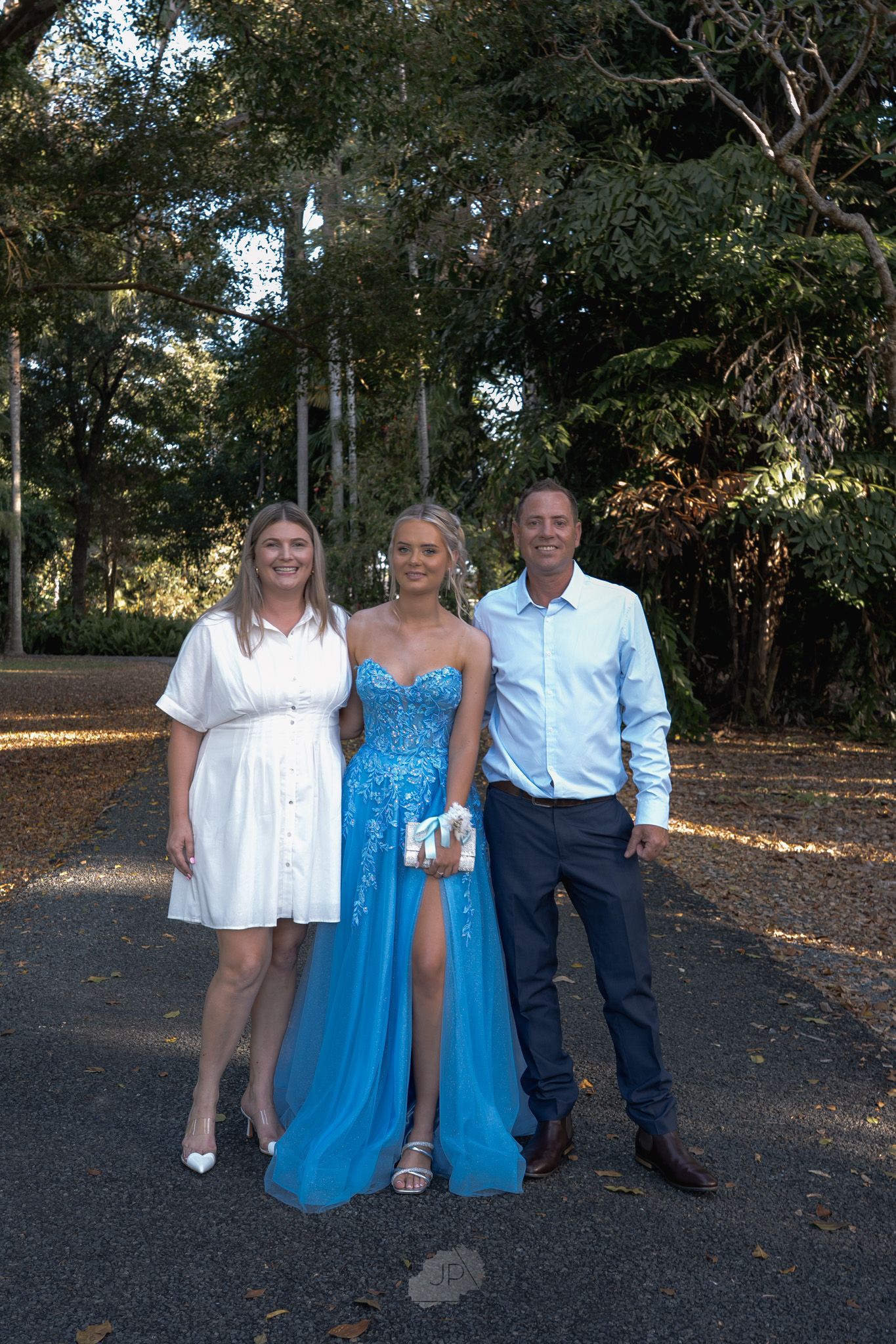 A group in white and blue outfits poses together outdoors on a shaded path surrounded by trees.