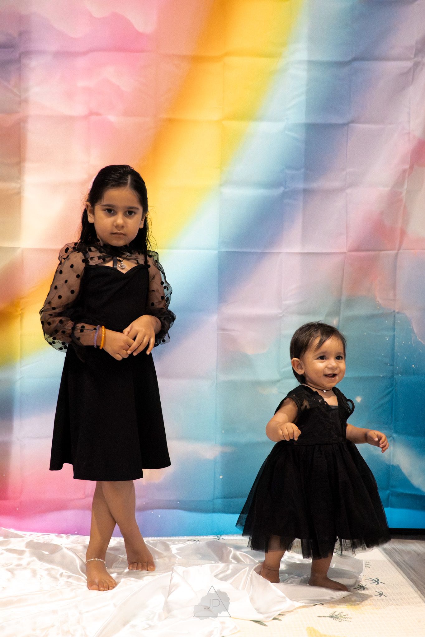 Children pose playfully against a colorful rainbow backdrop with soft white fabric on the floor during a photo session.