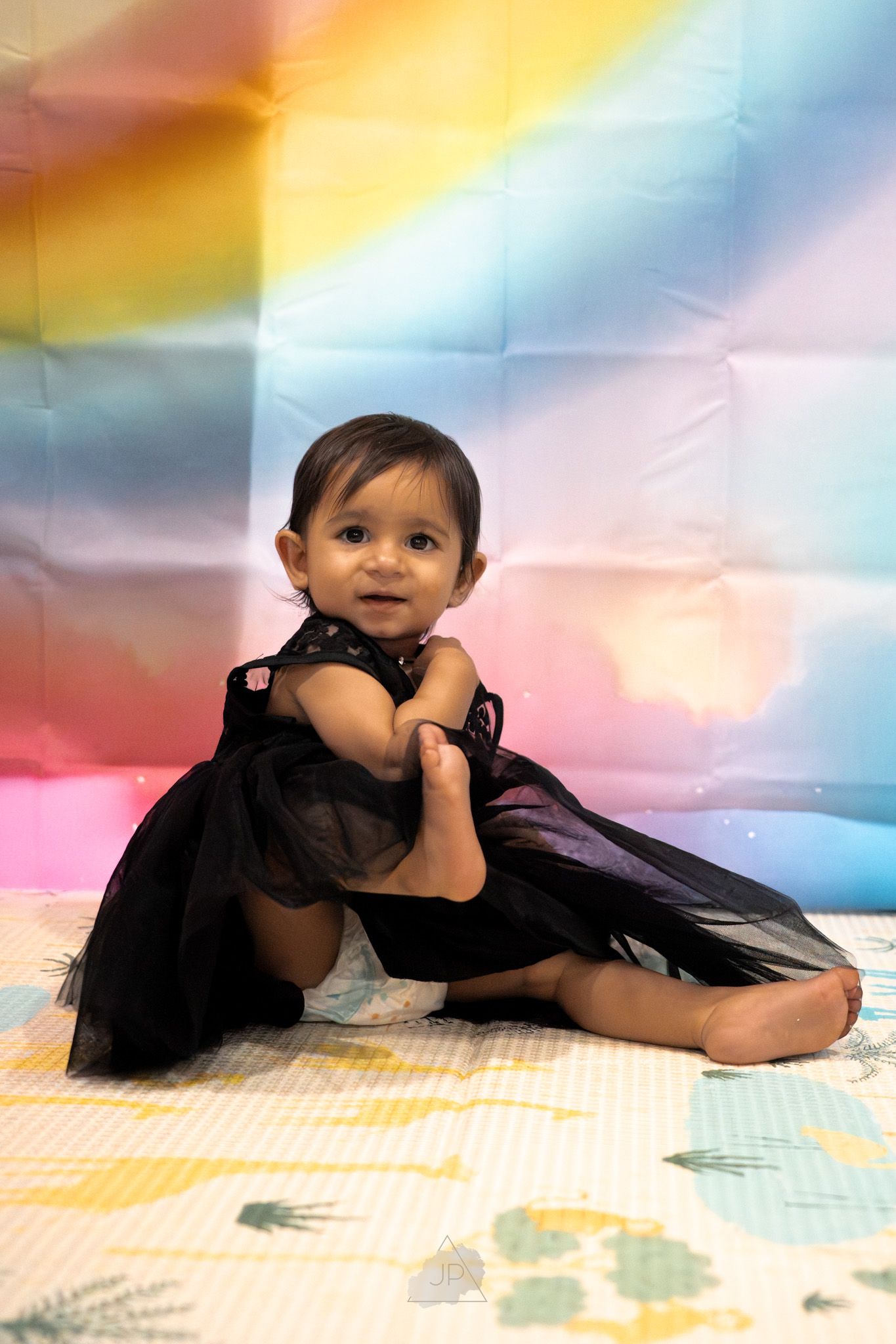 A small child in a black tulle dress sits on a colorful backdrop with pastel rainbow colors.